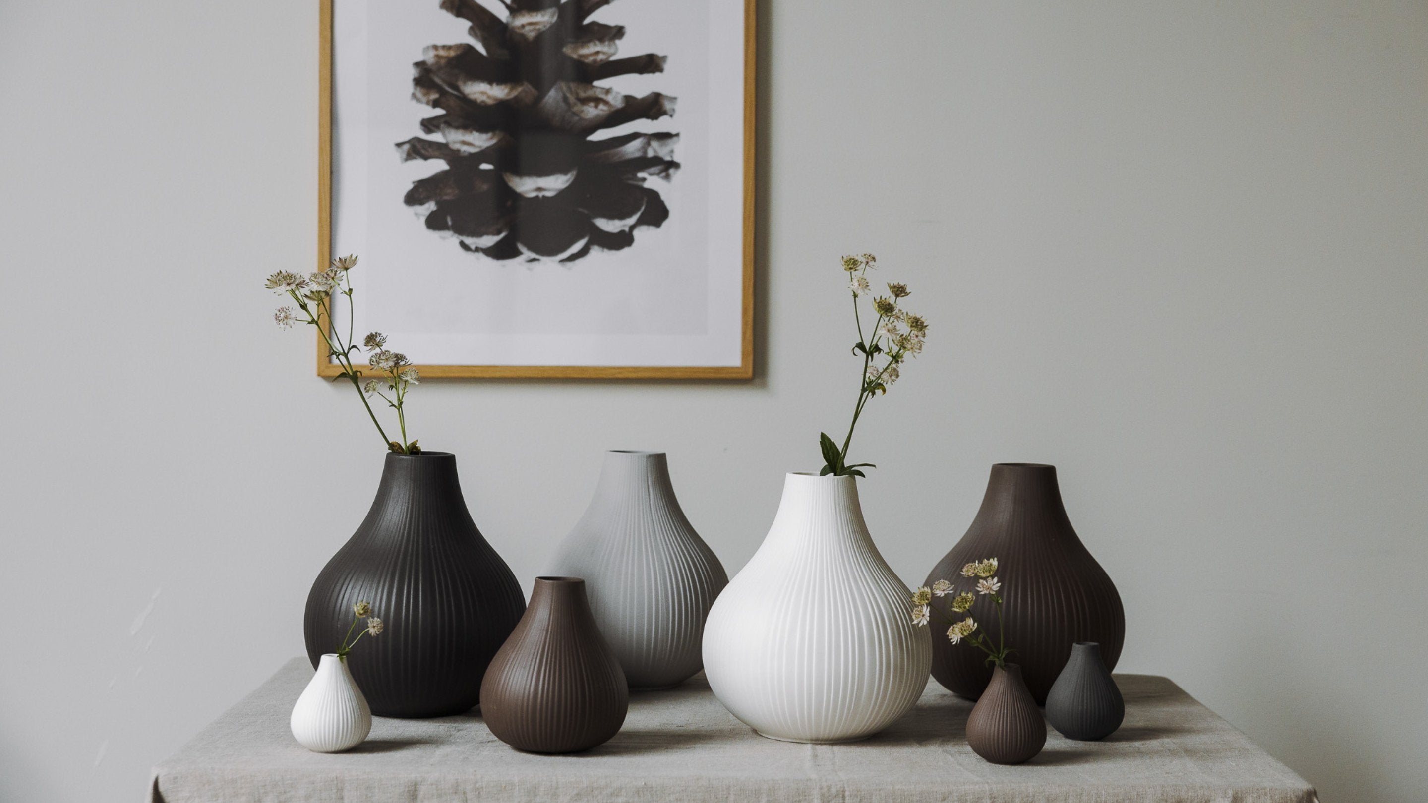 Ett arrangemang av vaser i jordnära toner står på ett bord, med en inramad bild av en tallkotte i bakgrunden.