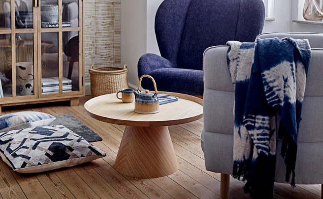 Ett mysigt vardagsrum med ett runt soffbord i trä, en tekanna med koppar, en blå fåtölj, en grå fåtölj draperad med en mönstrad pläd, golvkuddar och ett skåp med vitrinskåp.