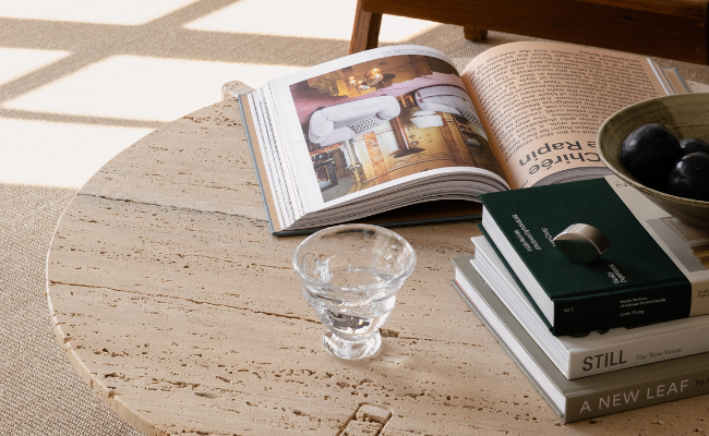 Stilren inredningsbild med ett soffbord i travertin, dekorerat med en uppslagen inredningsbok, coffee table books,, en glaskopp med vatten, en skål med mörka frukter och en stapel eleganta coffeetable-böcker.