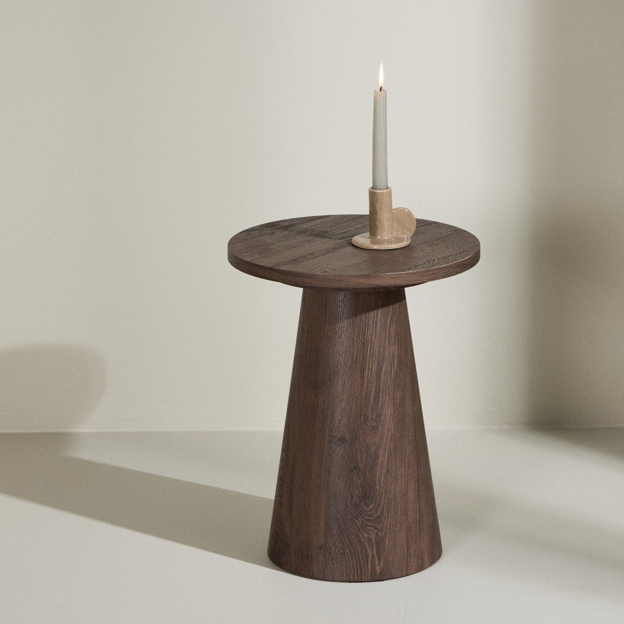 Lanzo Sidobord från Venture Home, med rund form och modern mocca-finish, står vid en ljus vägg. Ovanpå den sitter ett tänt vitt ljus i en beige hållare – en tidlös design.