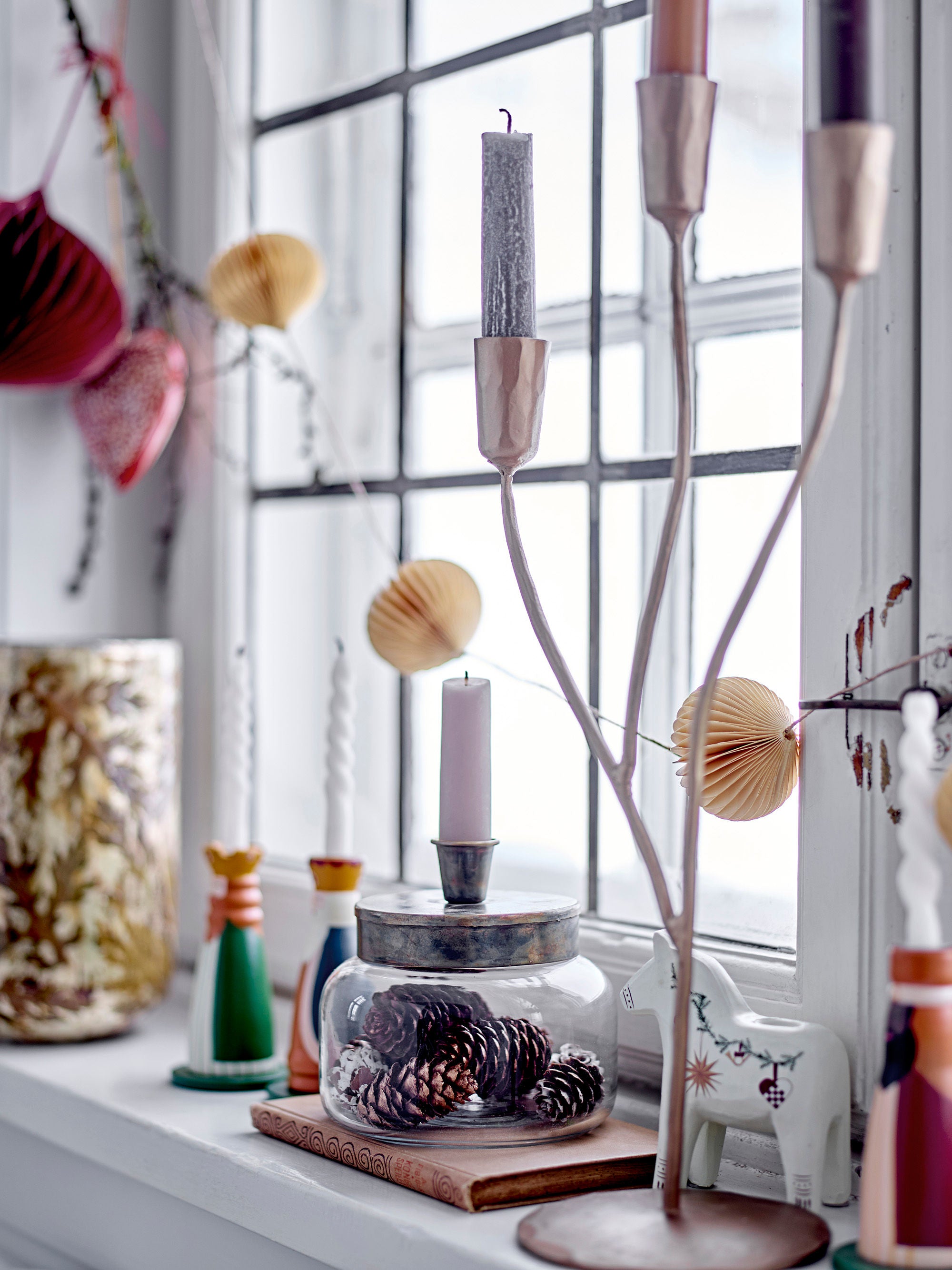En fönsterbräda prydd med levande ljus, en glasburk med kottar, en vit dekorativ häst, pappersprydnader och naturligt ljus som skiner genom glasrutor får en elegant accent av Bloomingville Lorine ljushållare i guldmetall.