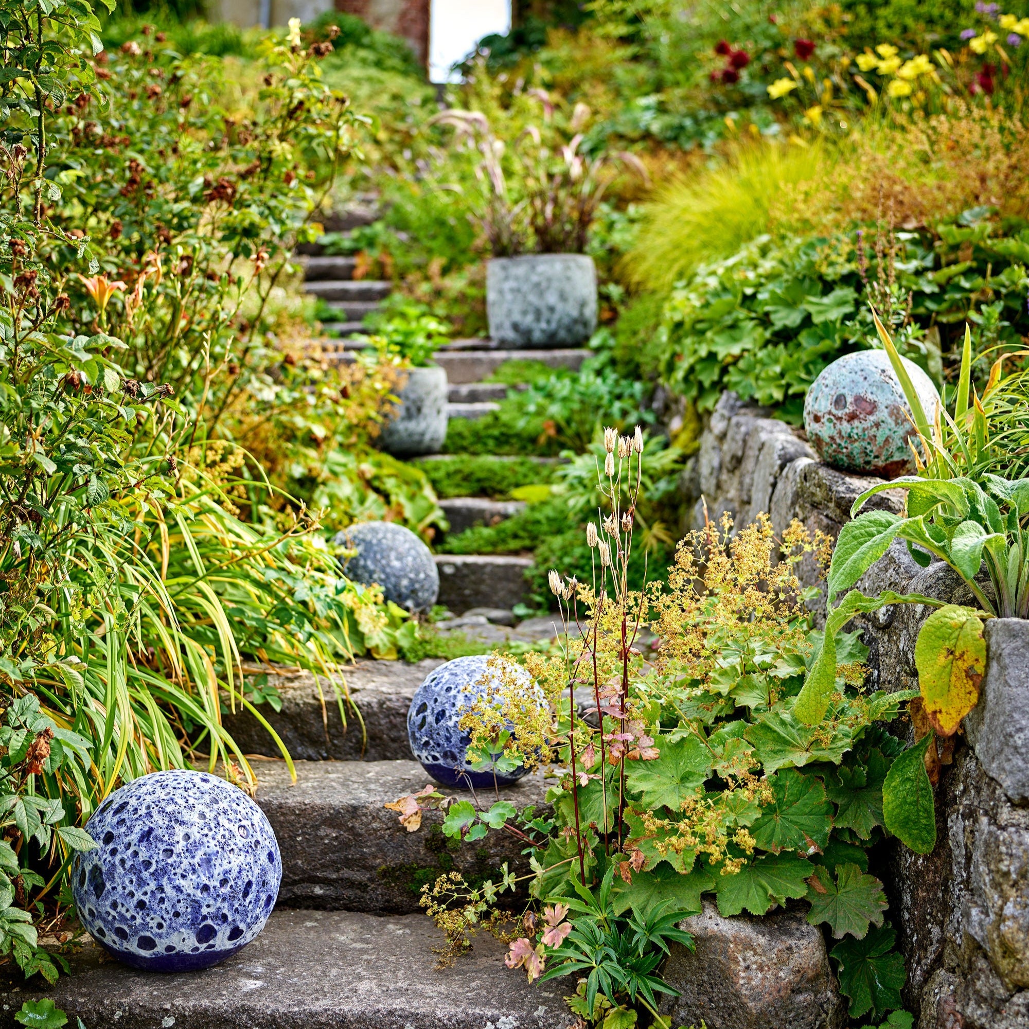 En lummig trädgård med en slingrande stenlagd trappa, omgiven av grönska och blommande växter. Dekorativa sfärer i blått och grönt pryder trappstegen och skapar en harmonisk och lekfull känsla i den naturliga miljön. I bakgrunden syns en rustik tegelbyggnad med en öppen dörr, vilket förstärker den inbjudande atmosfären.