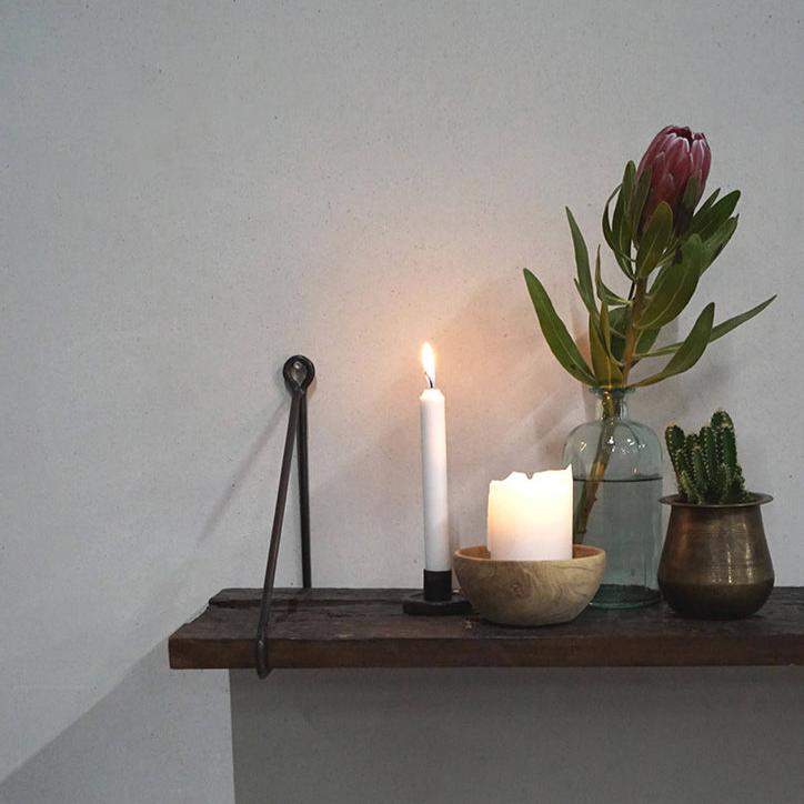 En Rustik Hylla i Återvunnet Trä med Svarta Fästen av Trademark Living håller ett tänt vitt ljus, en liten träskål med ett annat ljus och en genomskinlig vas med en rosa proteablomma. En mässingsbehållare rymmer en kaktus, medan den enkla väggen förstärker hyllans rustika charm.