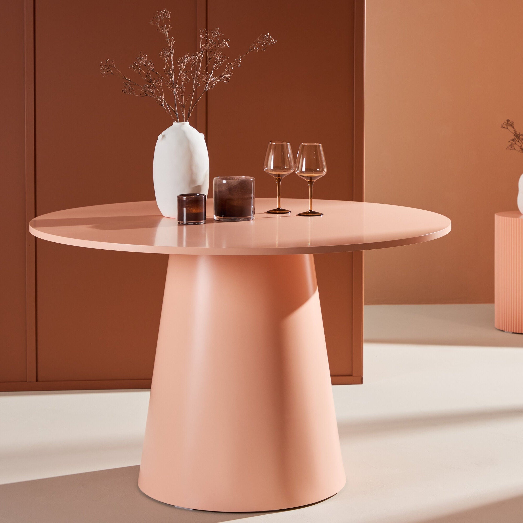 Venture Home Lanzo Matbord ⌀120 i persika pryds av en vit vas med torra grenar, två bruna glas och ett ljus. En matchande liten piedestal i bakgrunden rymmer ytterligare en vit vas med grenar.