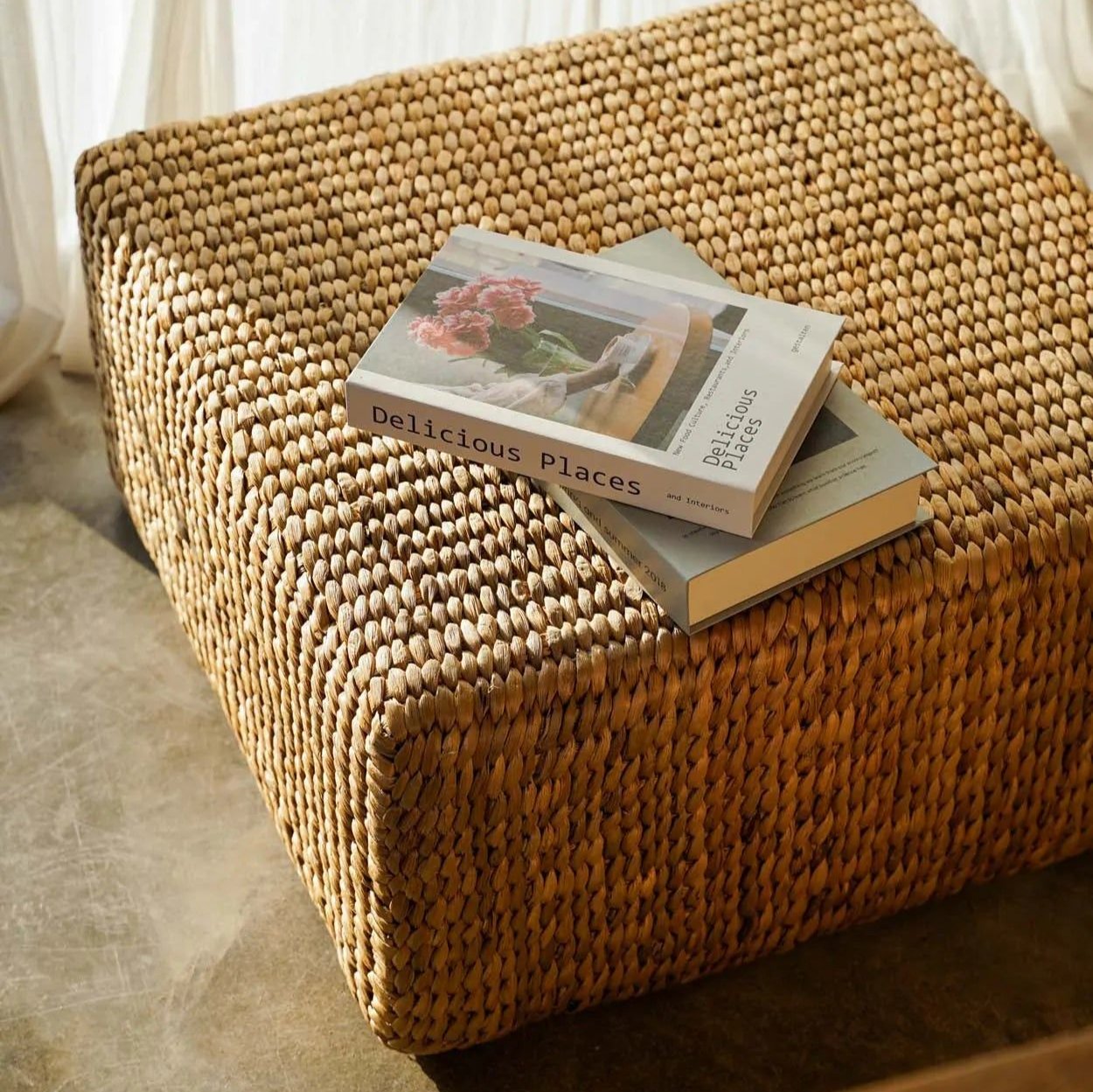 Två exemplar av "Delicious Places" är staplade på en Fyrkantig Puff i Vattenhyacint av bazar bizar, gjord av texturerat vävt material. Solljuset strömmar genom fönstret och skapar en varm och inbjudande atmosfär.