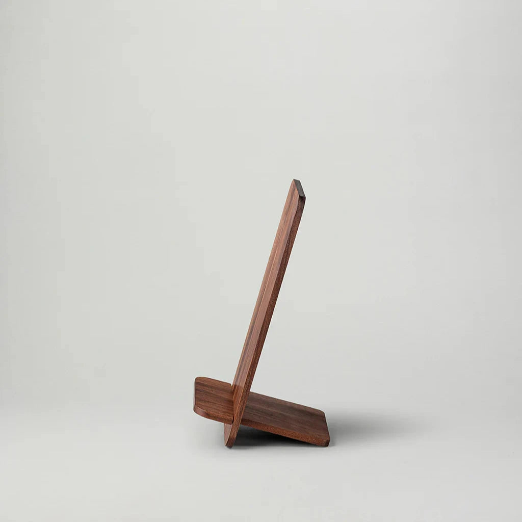NM Bokställ – Valnöt från New Mags är ett minimalistiskt designbokställ tillverkat av mörkbrunt trä med valnötsfinish. Dess eleganta, enkla design har två sammankopplade delar som bildar ett vinklat stöd. Den enkla, ljusa bakgrunden accentuerar monterns rena linjer och hantverk.