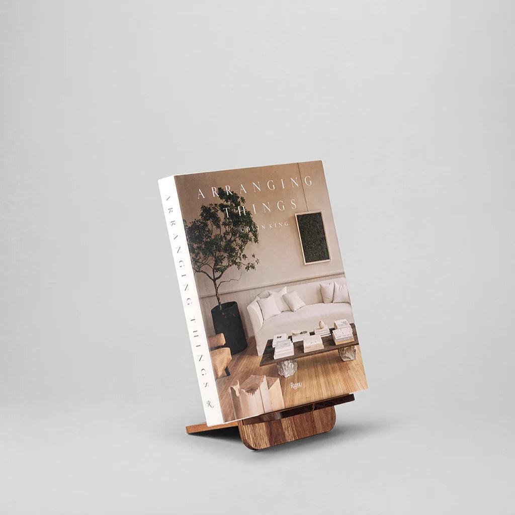 En bok med titeln "Arrangera saker" visas på NM Bokställ – Valnöt av New Mags. Stativet har en rik valnötsfinish, och bokens omslag visar upp ett minimalistiskt vardagsrum med en soffa, soffbord och inomhusväxt mot en vanlig ljusgrå bakgrund.