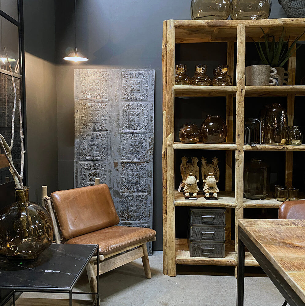 Ett mysigt rum med en trähylla, bruna glasföremål, inredning, James Loungefåtölj i Brun Läder med Mangoträ från Trademark Living, rustikt metallskåp, statyer, stor glasvas och utsmyckad grå metalldörr mot mörka väggar.