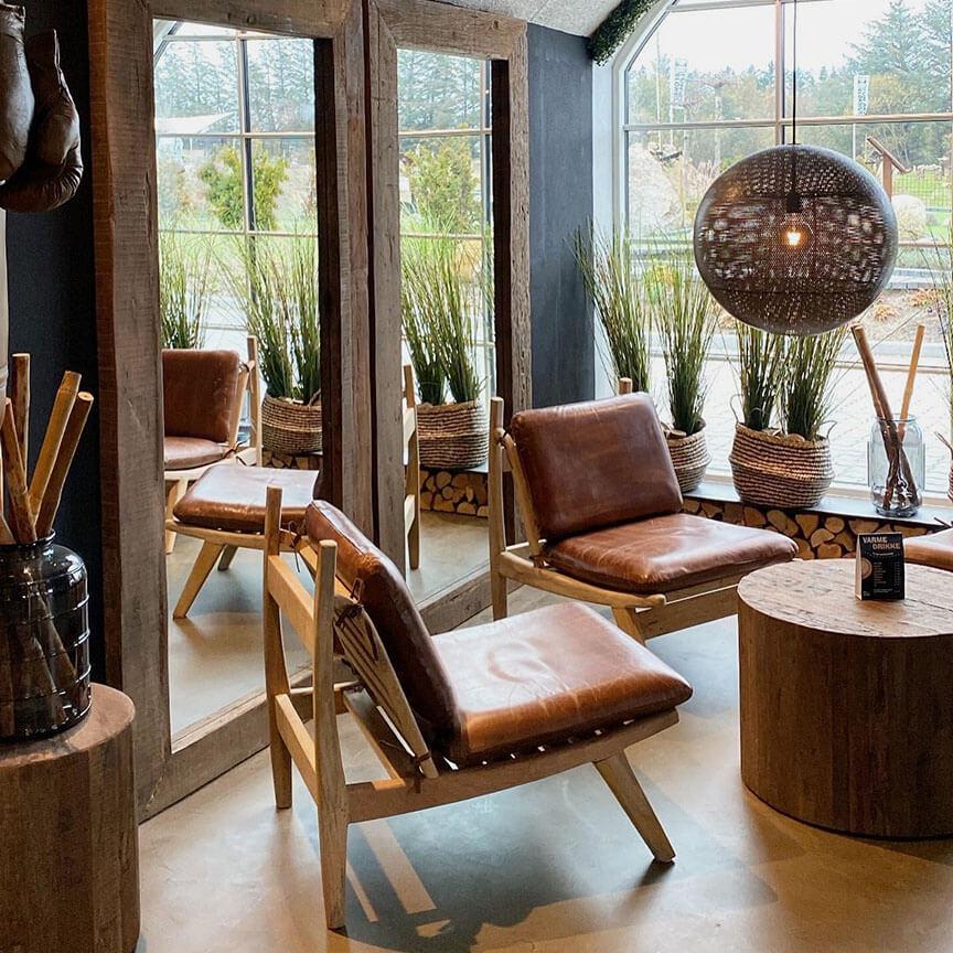 Mysig modern lounge med James loungefåtölj i brun läder med mangoträ från Trademark Living, ett runt bord i massivt mangoträ, stora speglar, flätade korgar med högt gräs och en globependel vid fönster med utsikt över träd.