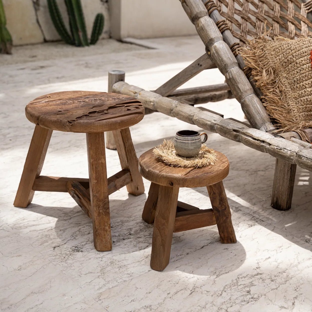 Två Yatai Pall pallar i Återvunnet Teakträ by bazar bizar sitter på en sten uteplats bredvid en flätad solstol. En pall rymmer en keramisk mugg på ett halmunderlägg, vilket framhäver elegant detalj. Gröna kaktusväxter kompletterar den naturliga miljön i bakgrunden.