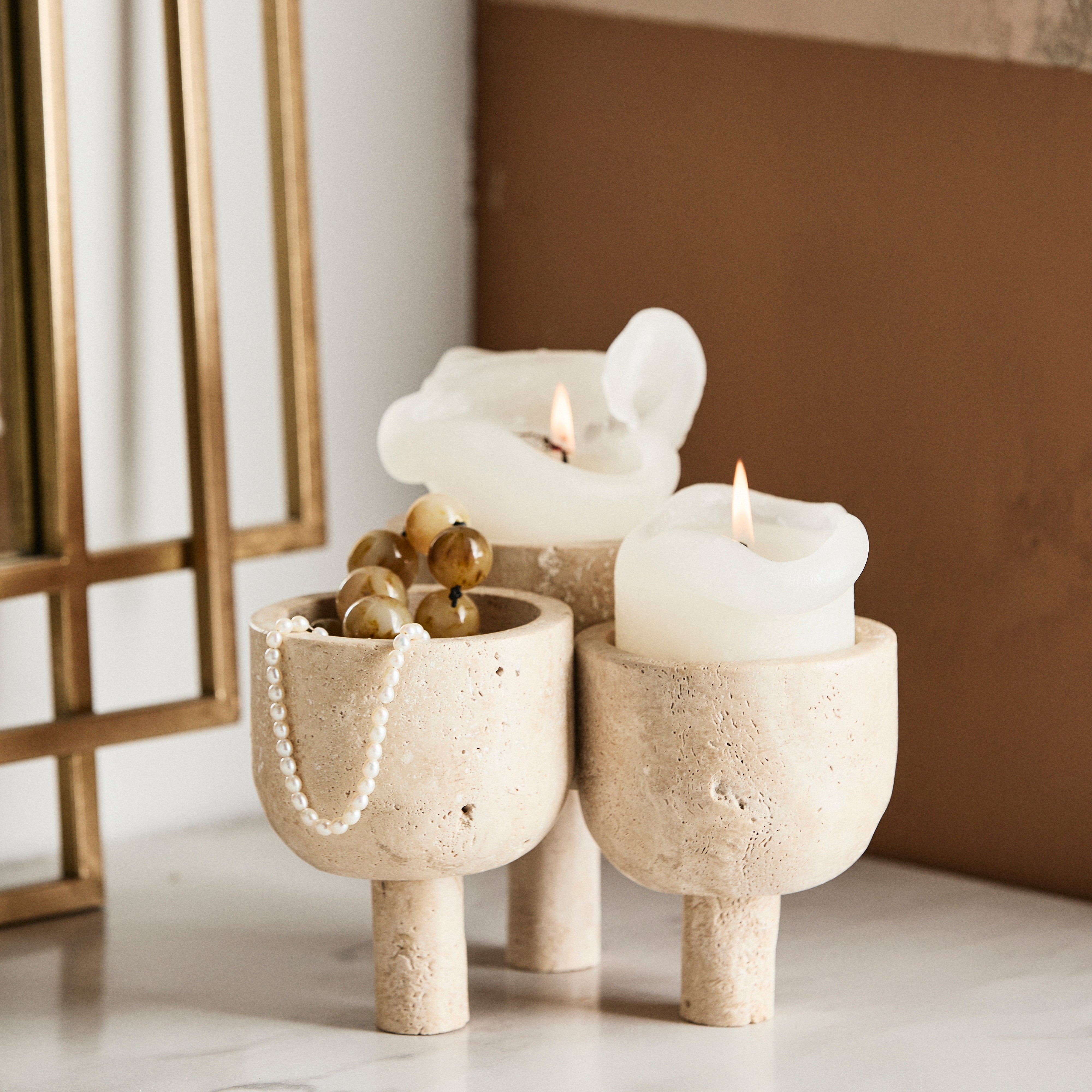 Tre ROCA - Skulptural Förvaring i Travertin-ljusstakar från Nordal, i beige travertin med korta ben, visar två tända vita ljus och ett pärlarmband på en vit yta bredvid en guldinramad spegel, vilket ger en charm av natursten.