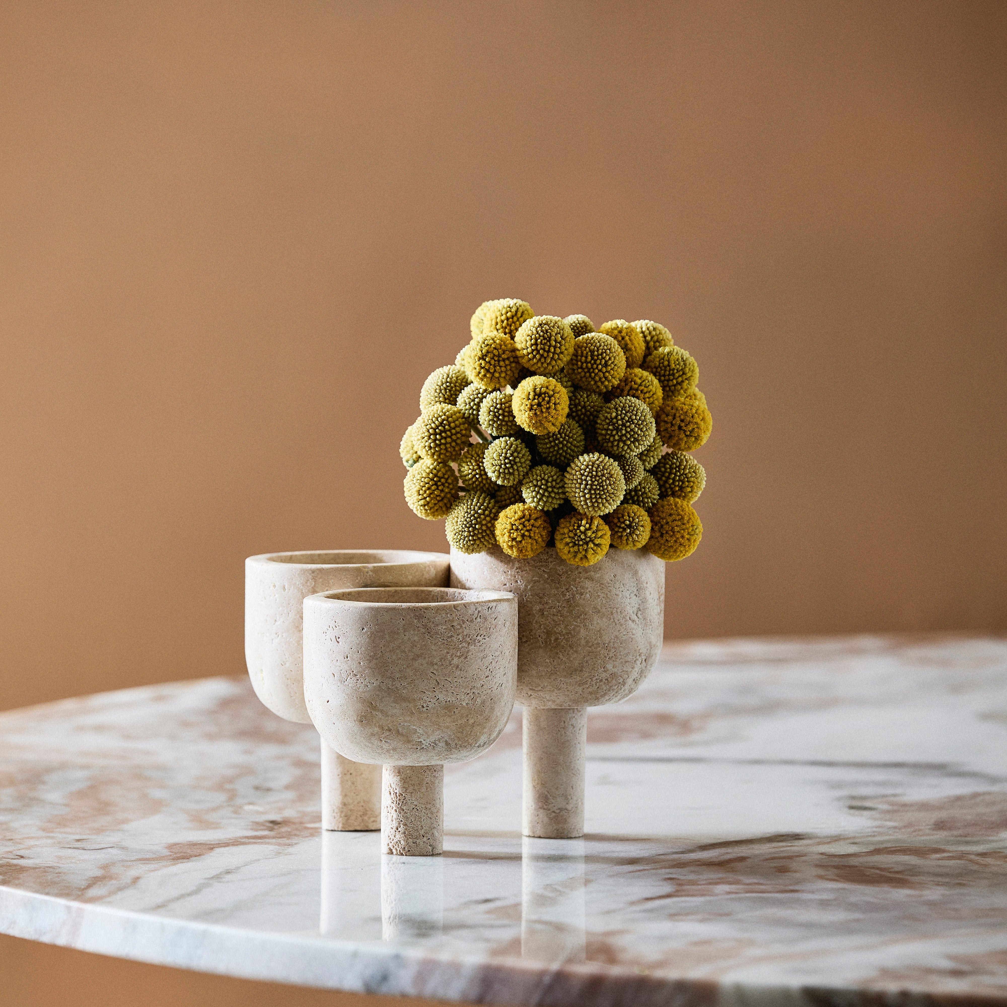 Tre beige keramikvaser med korta, cylindriska baser från Nordal står på ROCA - Skulptural Förvaring i Travertin; i en vas finns små gula blommor mot en varm brun bakgrund.