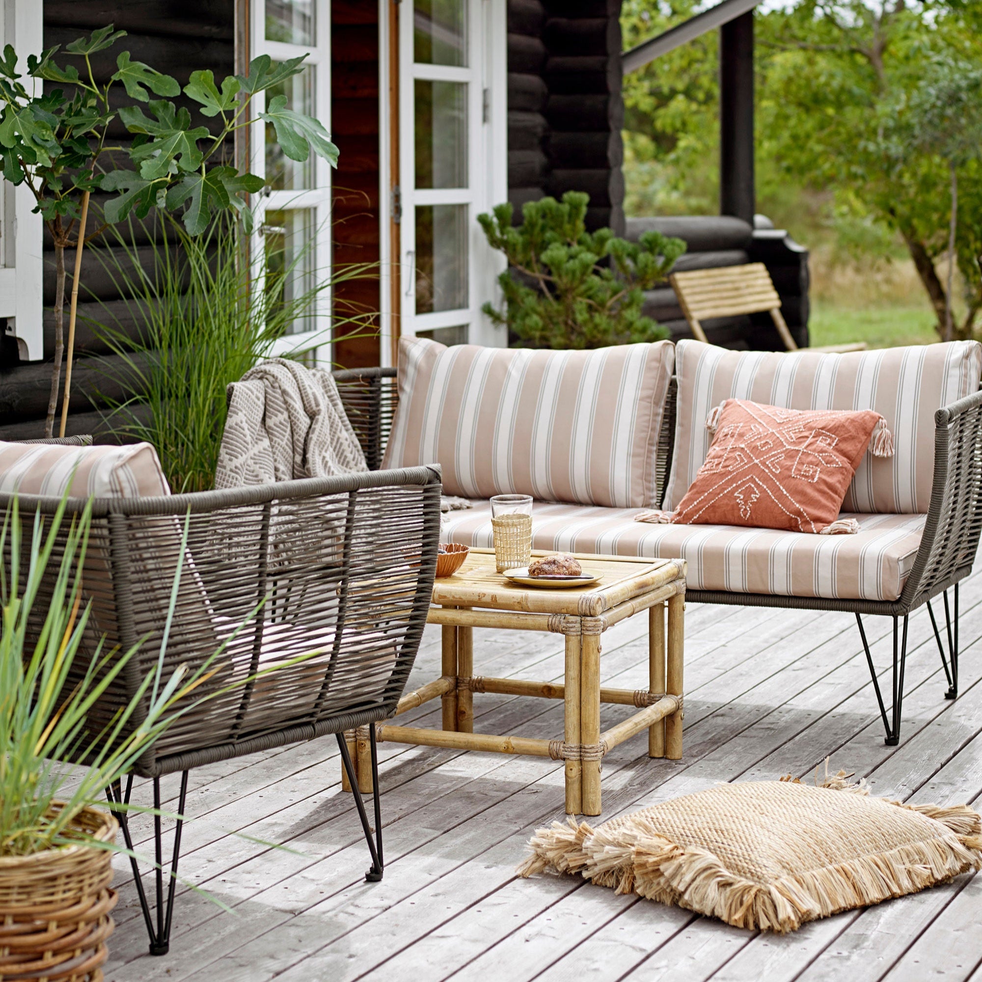 En uteplatsscen visar upp en Mundo Utesoffa från Bloomingville med flätat och handvävda detaljer, kompletterat med matchande stolar och randiga kuddar. Ett träbord visar glas och dekor, omgivet av gröna växter, vilket förstärker den hållbara designen. Ett trähus syns delvis i bakgrunden.