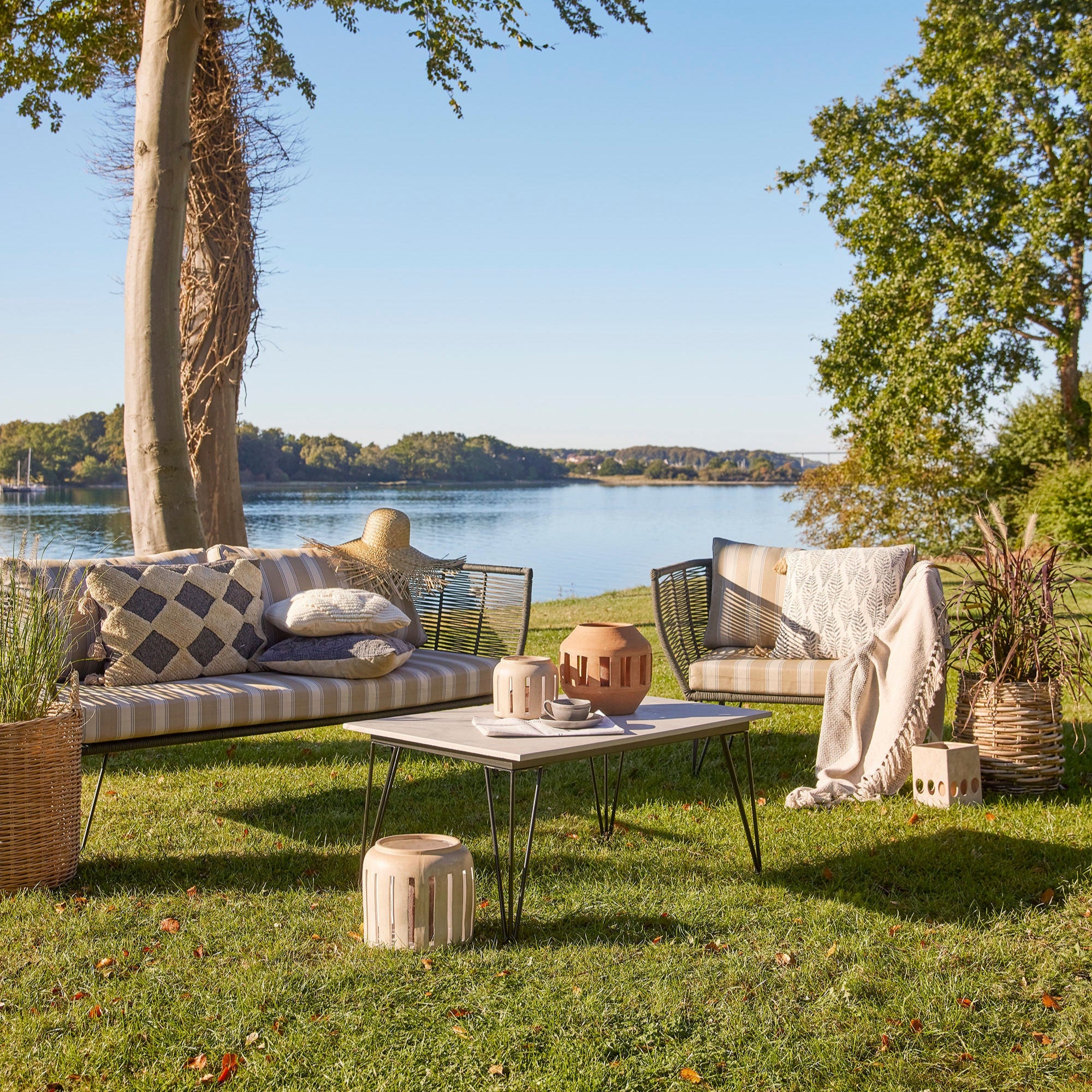 En uteservering vid en sjö har två Bloomingville Mundo Utesoffa-bänkar, ett soffbord och diverse kuddar. En stråhatt står på ena bänken, omgiven av krukväxter och dekorativa lyktor. Träd och lugnt vatten förstärker den moderna designen under en klarblå himmel.