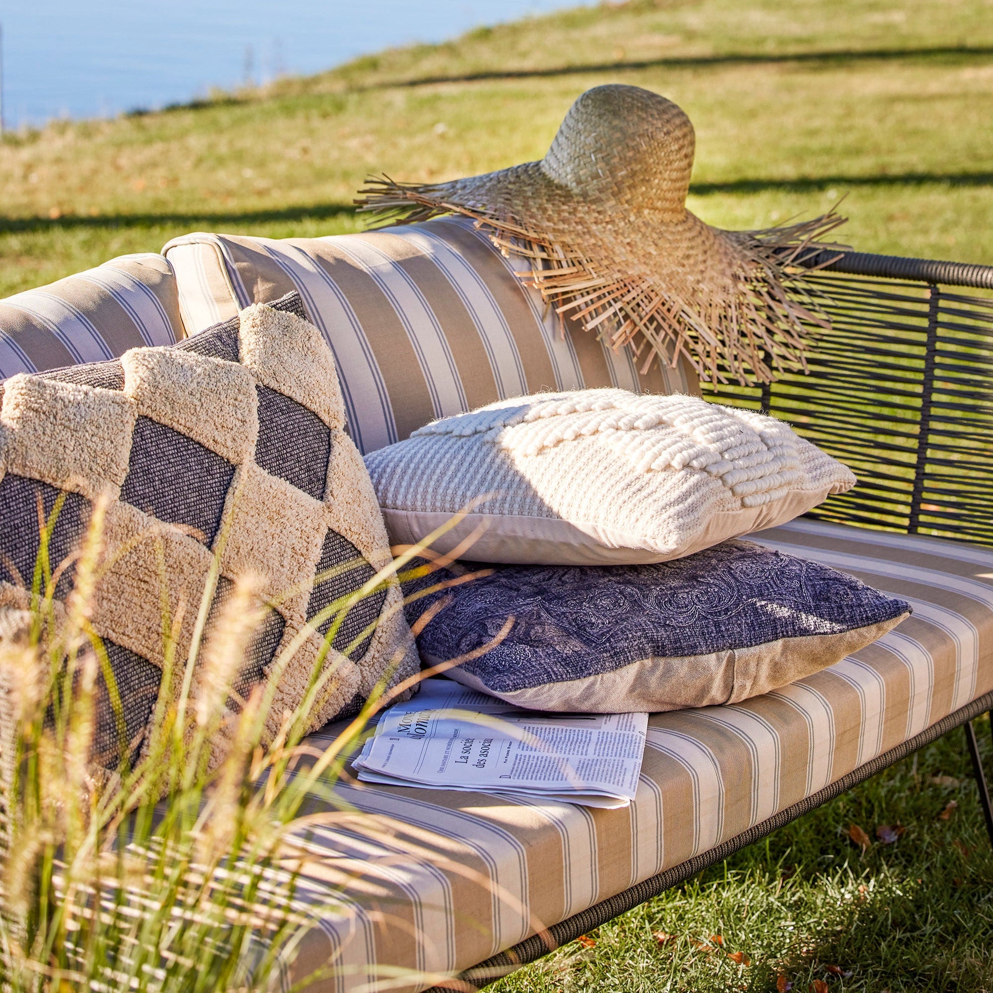 Mundo Utesoffa från Bloomingville, en snygg trädgårdssoffa med handvävda detaljer i grönt och metall, har rutiga och texturerade kuddar, en tidning och en stråhatt på gräs med utsikt över vattnet. Den gräsbevuxna förgrunden ger djup till den lugna scenen.