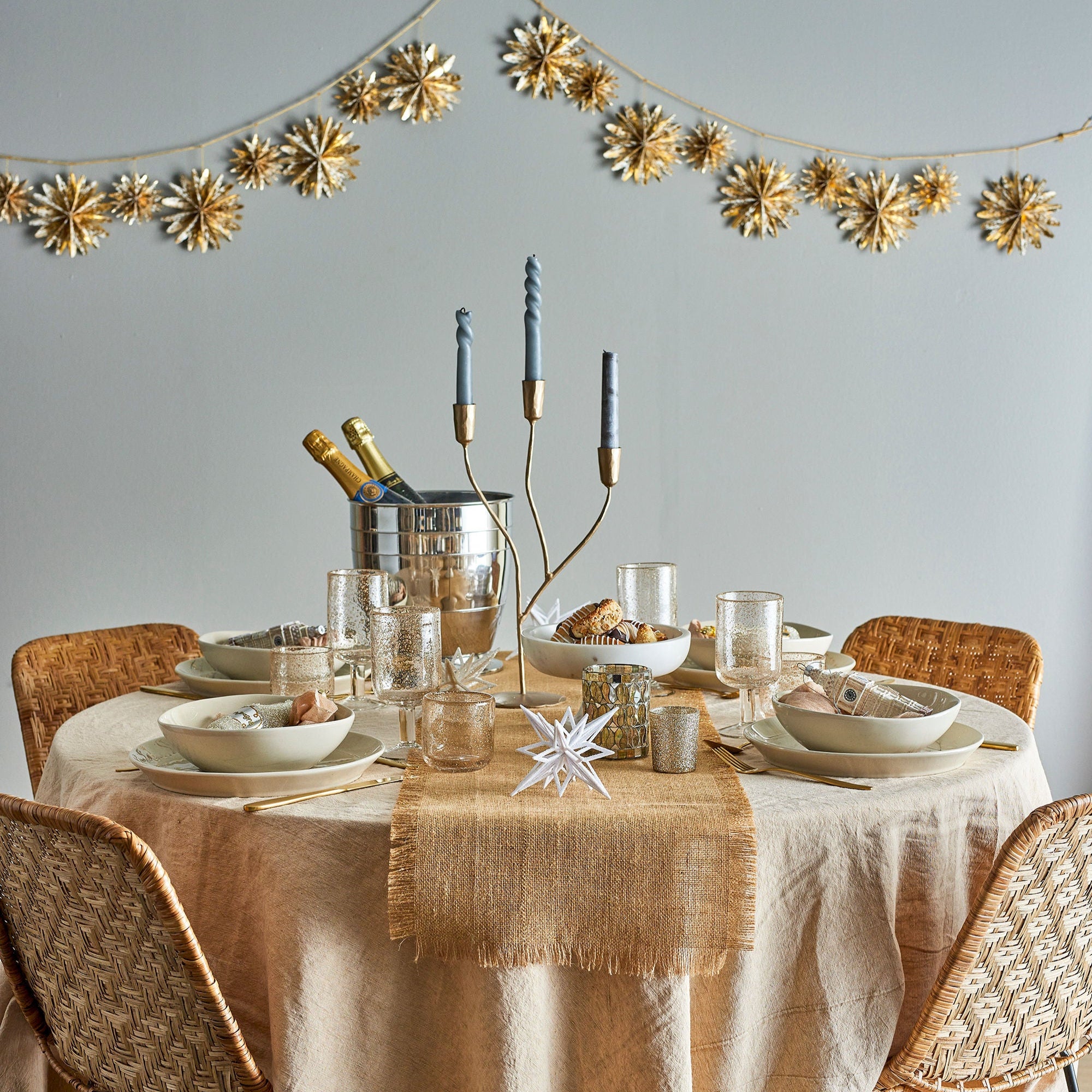 Ett runt matbord för fyra personer med neutrala tallrikar, glas och servetter. Guldaccenterna inkluderar en Bloomingville Lorine Candle Holder, en champagnehink och en girlang med guldstjärndekorationer på väggen. Fyra vävda stolar omger bordet.