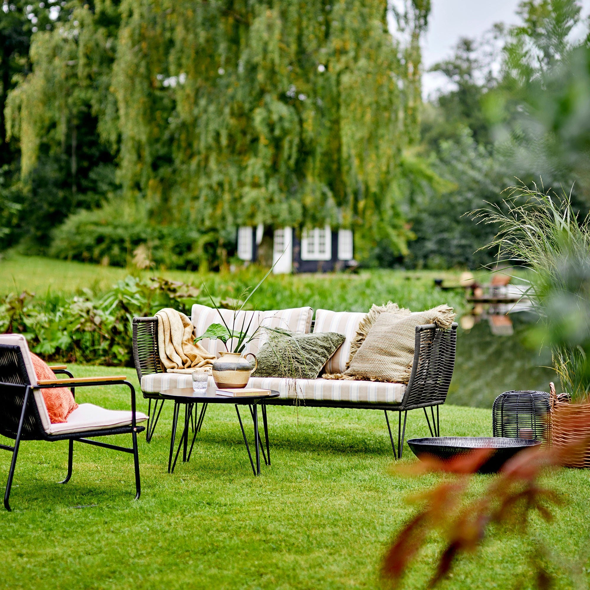 Uteserveringen i en lummig trädgård har Mundo Utesoffa från Bloomingville - en elegant trädgårdssoffa med handvävda gröna och metalldetaljer - tillsammans med en fåtölj, bord, dekorativa kuddar och en växt. En damm och träd skapar en lugn bakgrund medan ett litet hus förstärker den moderna designen.