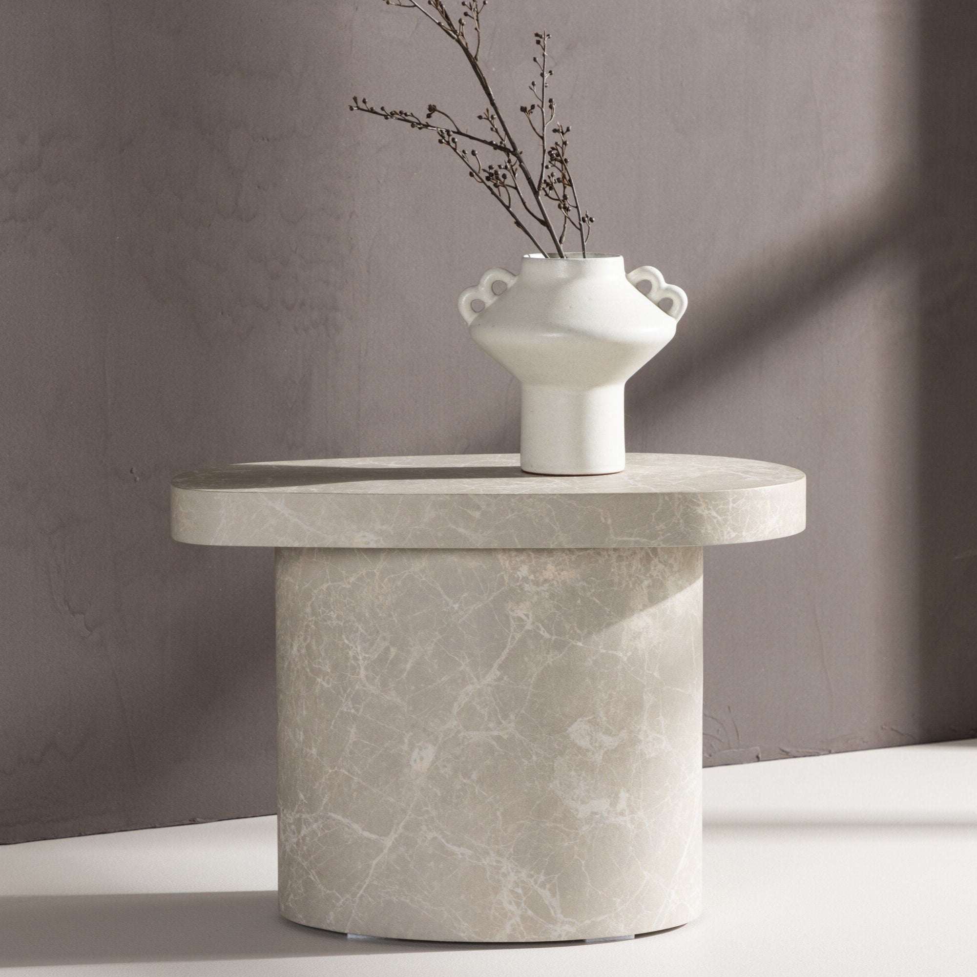 Venture Home Båstad Sidobord, Concrete Look (30x50 cm), med sin runda topp och cylindriska bas, står mot en ljusgrå vägg. En vit keramikvas med handtag och torkade grenar sitter på toppen, perfekt för minimalistisk inredning.