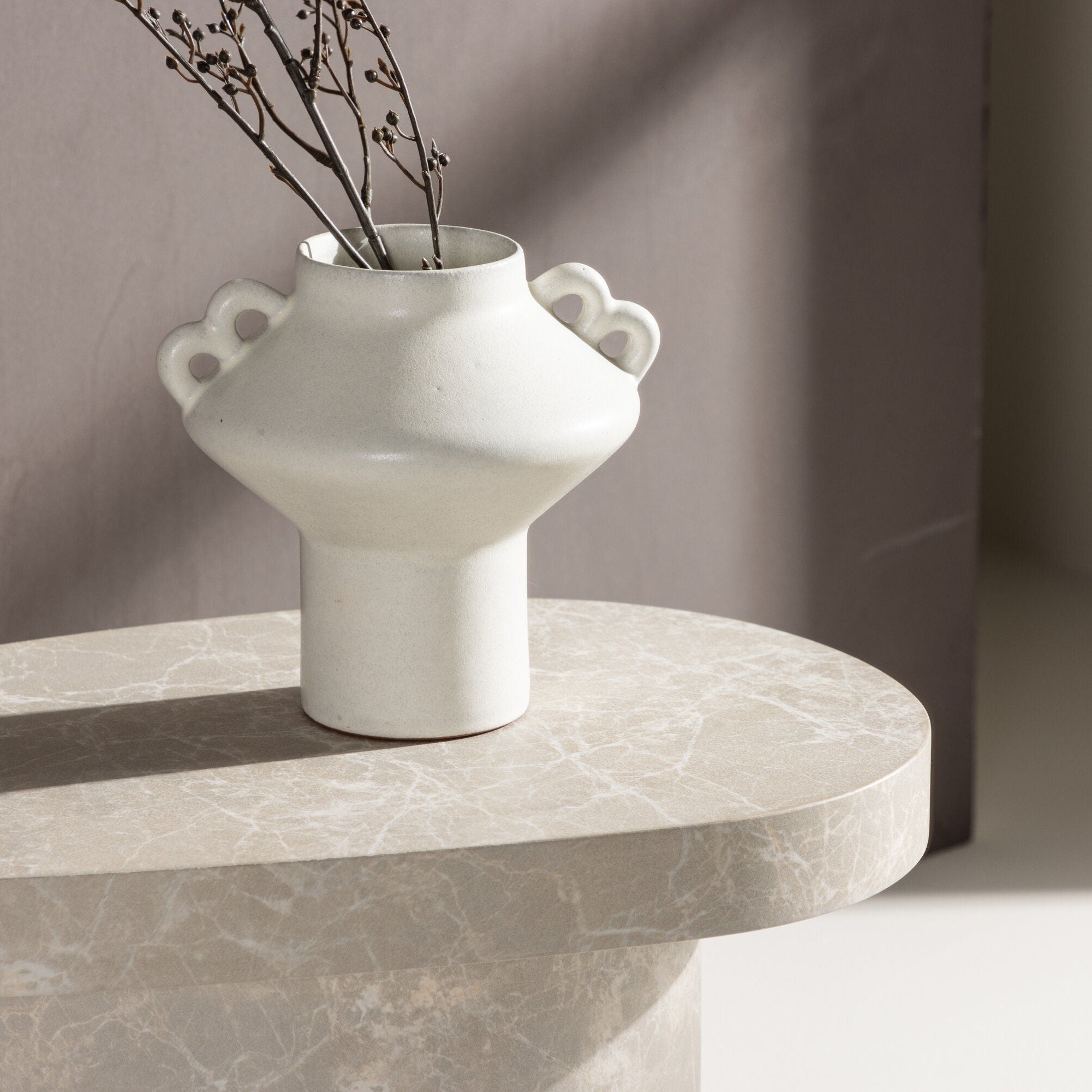 En vit keramikvas med två handtag som håller i tunna grenar står på Venture Home Båstad Sidobord, Concrete Look (30x50 cm), och det mjuka solljuset kastar skuggor - perfekt för minimalistisk inredning.