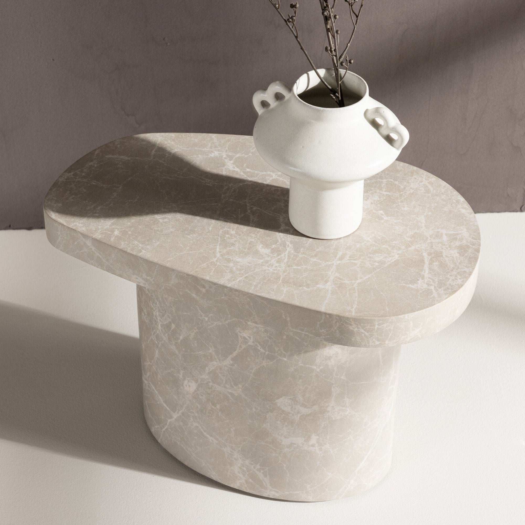 En minimalistisk, ovalformad Båstad Sidobord från Venture Home i betonglook (30x50 cm) har en vit keramikvas med handtag som håller i tunna grenar. Ställd mot en mjukt upplyst, enfärgad bakgrund - perfekt för minimalistiska interiörer.