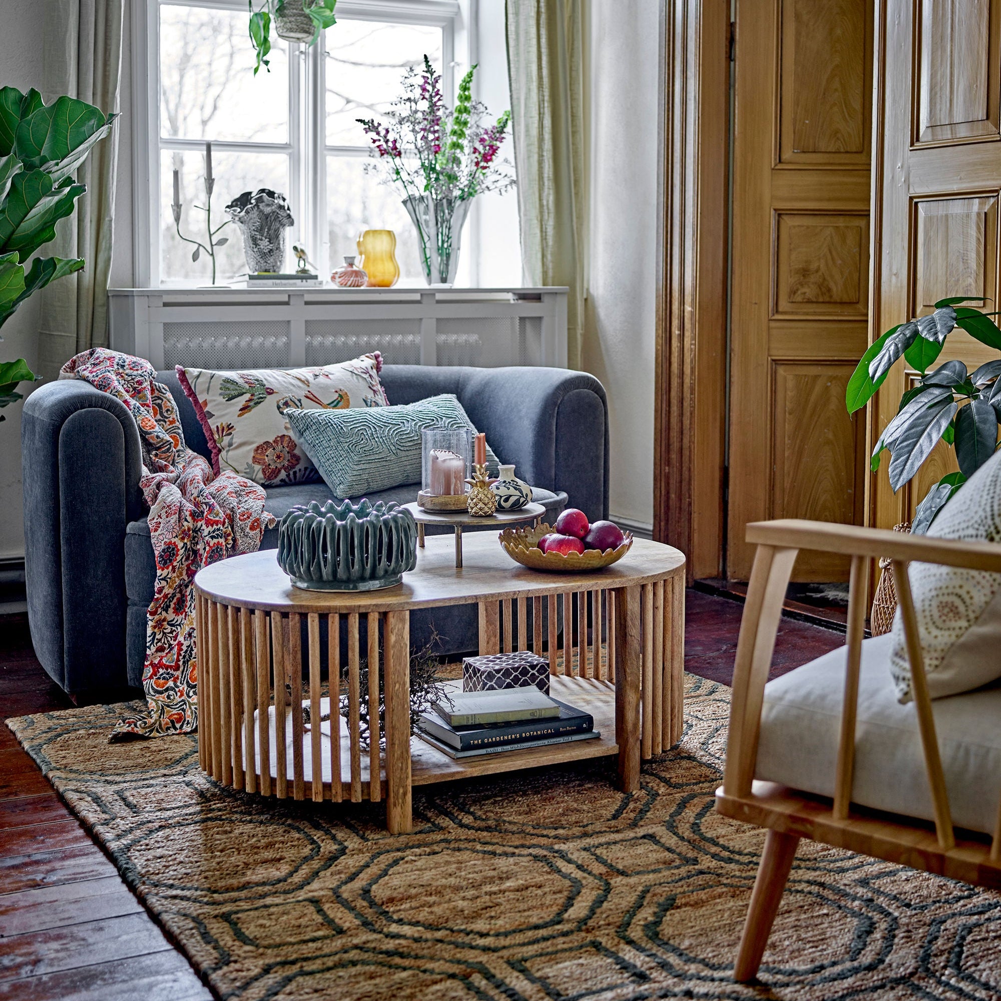 Ett mysigt vardagsrum har en grå soffa med färgglada kuddar och en pläd, Creative Collection Sali Soffbord – oval design i mörk mangoträ i mitten, en mönstrad matta, växter och skira gardiner som släpper in naturligt ljus för skandinavisk design.