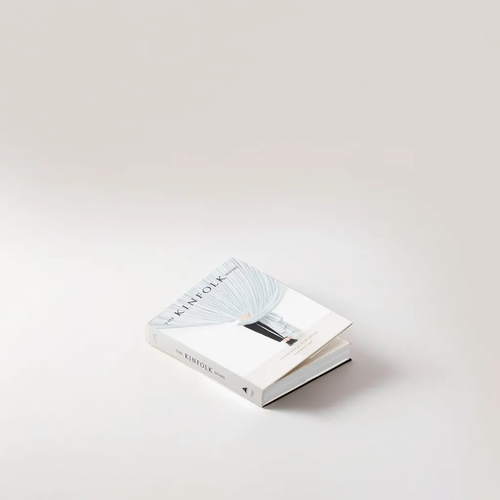 The Kinfolk Home" av New Mags ligger öppet på en minimalistisk, ljus yta, med en konstnärlig omslagsbild som återspeglar livsstil och design, som framhäver dess djupgående utforskning av hemmets estetik.