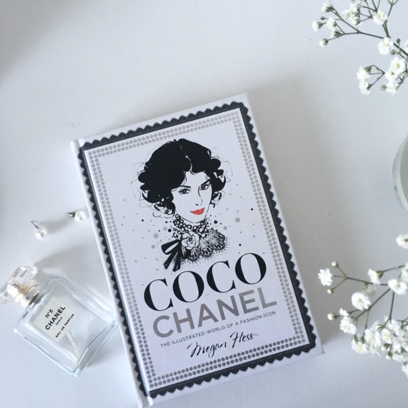 Den nya Mags-boken "Coco Chanel – The Illustrated World of a Fashion Icon" vilar elegant på en vit yta bredvid en Chanel parfymflaska, förstärkt av fina vita blommor och pärlörhängen, vilket gör den perfekt för alla modeälskare.