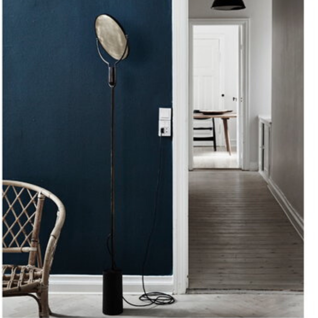 Ett minimalistiskt rum har en vävd stol, en hög golvlampa och en mörkblå vägg, som fångar den fridfulla essensen av The Kinfolk Home av New Mags. En vit dörr öppnar sig till en trägolvad hall som leder till ett bord och stolar i fjärran, vilket erbjuder enkelhet rik på detaljer.