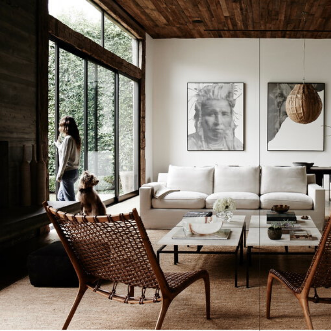 Det här mysiga vardagsrummet är inspirerat av The Kinfolk Home av New Mags och har ett trätak och stora fönster, med en vit soffa, vävda stolar med en värd för en slappande hund, ett soffbord i trä och abstrakt väggkonst. En person står vid fönstret och tittar ut.