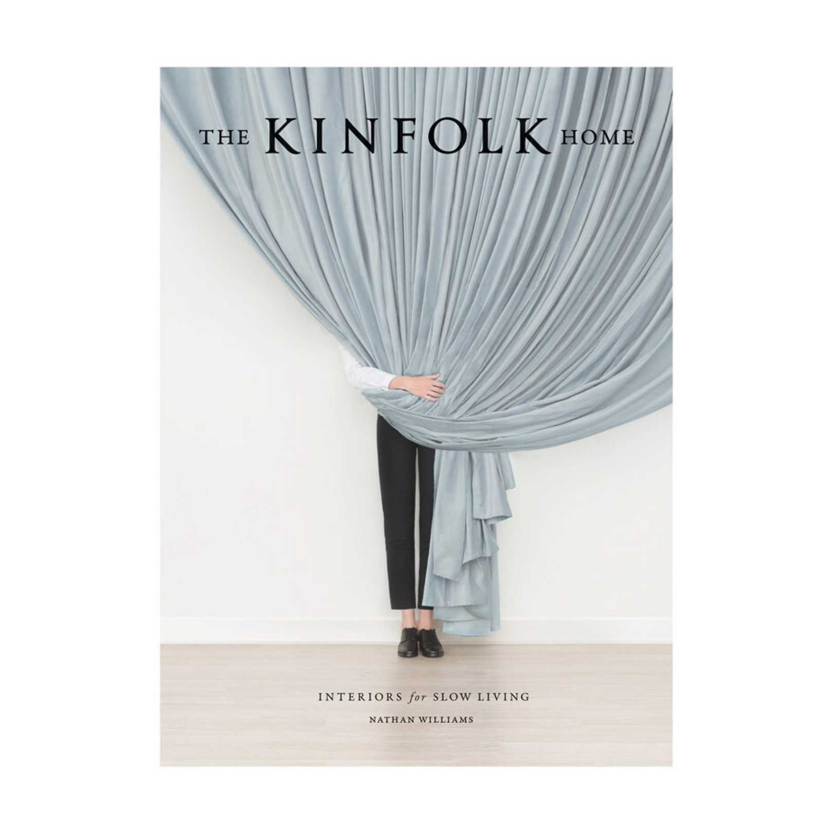 En person står bakom en stor draperad blå gardin, med bara fötterna och en del av ena armen synliga. Omslaget till "The Kinfolk Home" av New Mags erbjuder inspiration för de som har en inredningsintresse.