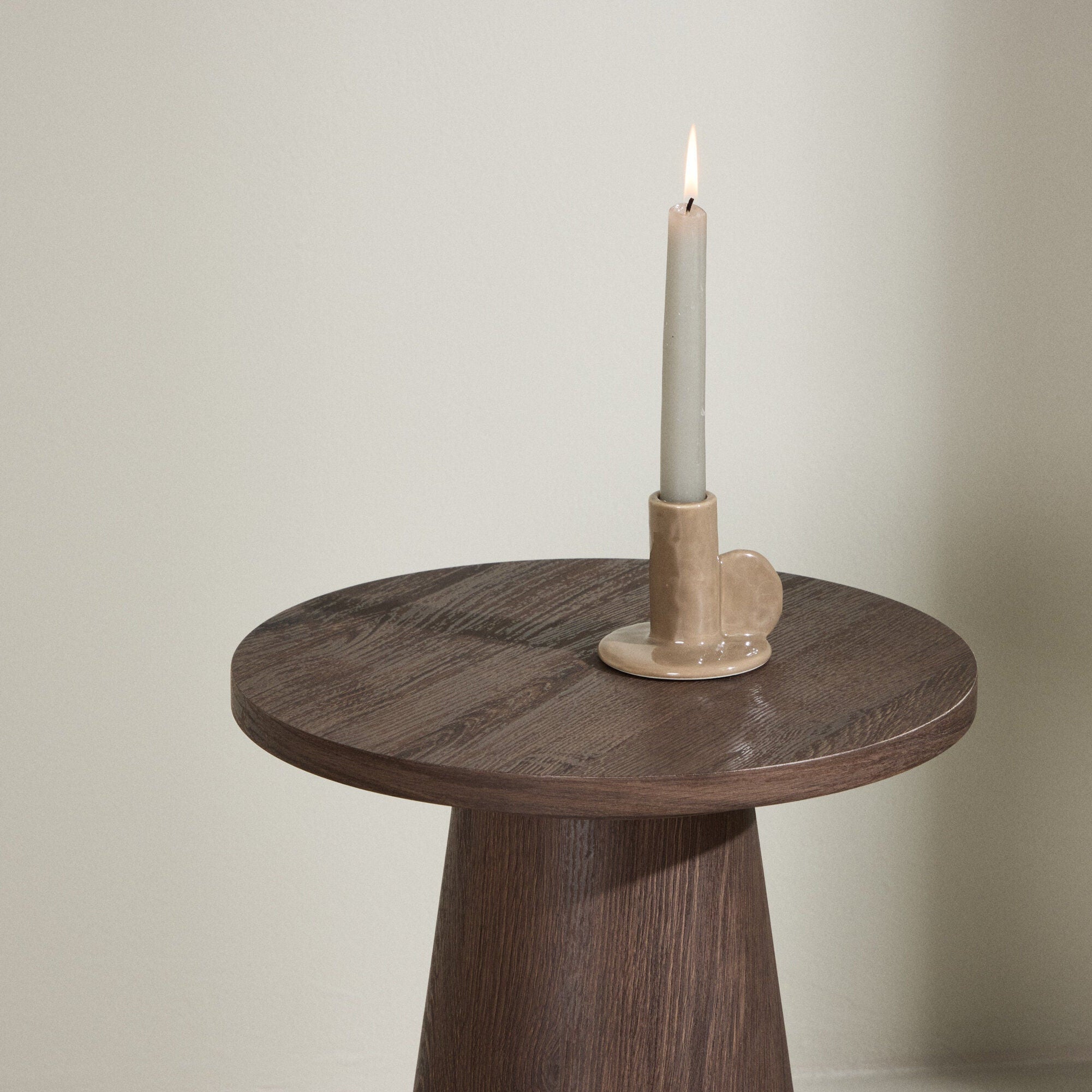 Ett tänt vitt ljus i en beige keramikhållare står på Lanzo Sidobord – Rund Form & Modern Mocca-Finish från Venture Home, vilket ger tidlös design till det kompakta, mörka bordet mot en ljus vägg.