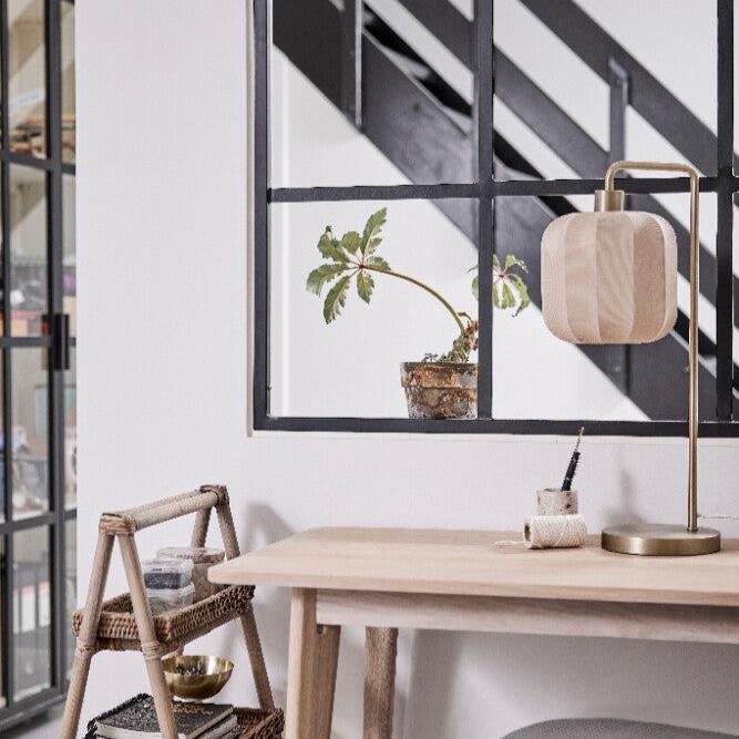 Bilden visar ett modernt, minimalistiskt skrivbord med en bordslampa och mässing till höger. På skrivbordet står även en liten behållare med pennor. Vid sidan av skrivbordet finns en liten stege i rotting med tre hyllor, varav en har en guldfärgad skål. I bakgrunden syns en inomhusvägg med svarta fönsterkarmar som skiljer två rum åt. På andra sidan av fönstret står en växt med gröna blad i en genomskinlig kruka. Svarta trappsteg och ett räcke syns också bakom glaset, vilket ger ett modernt industriell