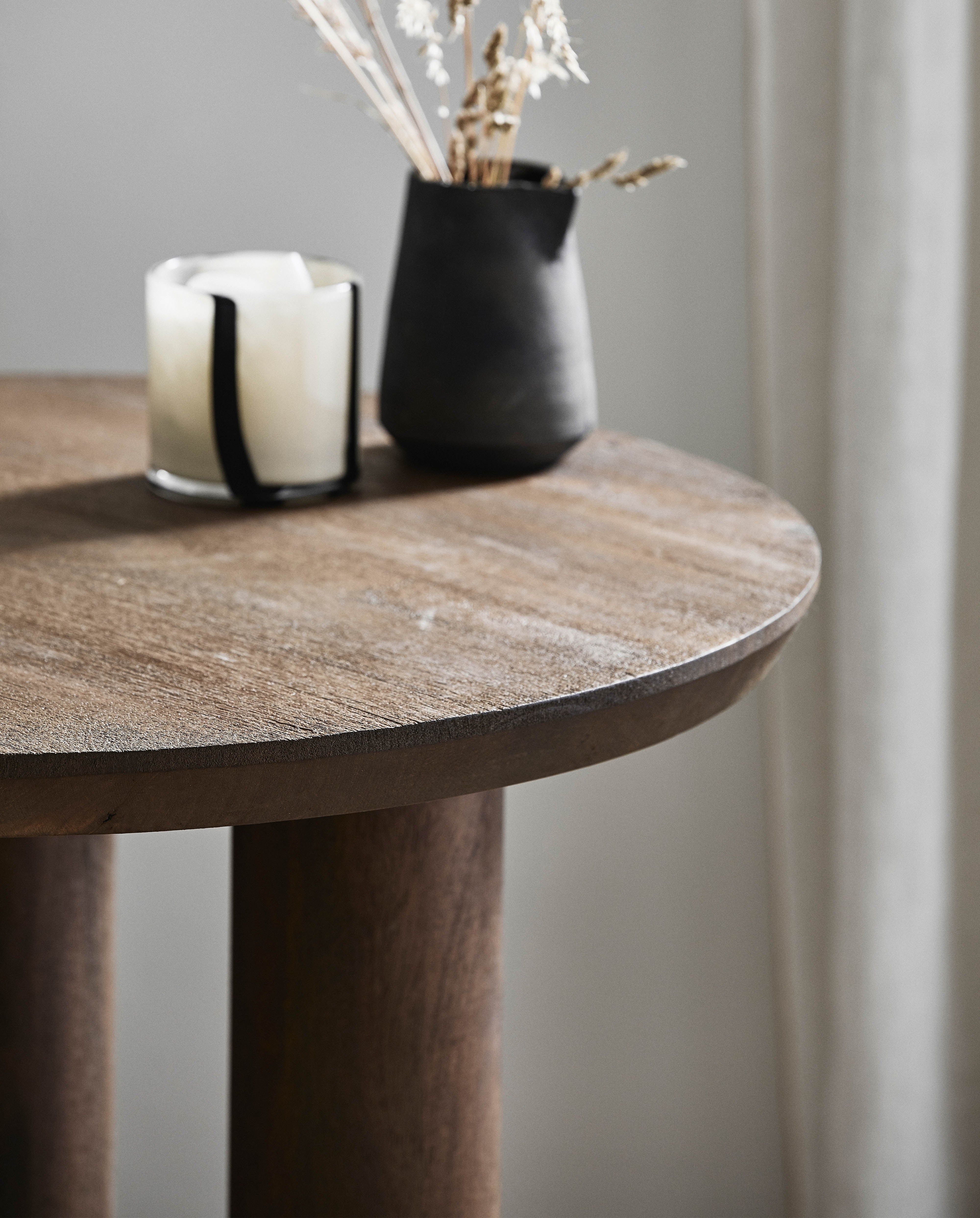 HELIN cafébord från Nordal i mörkt mangoträ med naturlig känsla, stilfullt dekorerat med vitt ljus i glaslykta och svart vas med torkat gräs mot en neutral bakgrund.