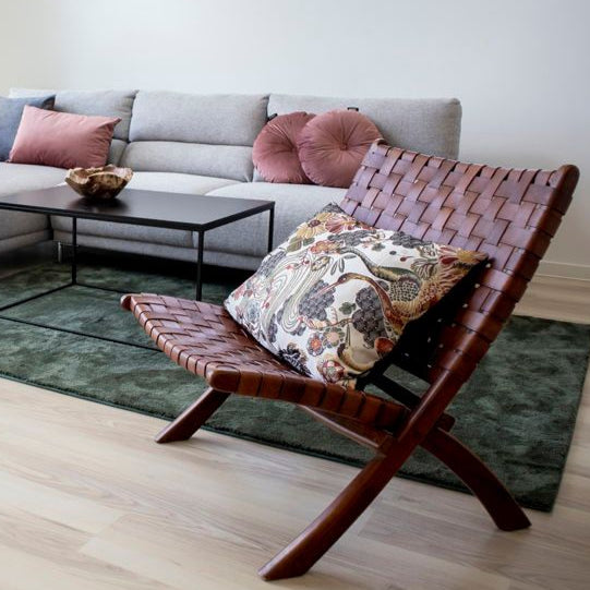Ett mysigt vardagsrum visar upp en Perugia Hopfällbar Stol från House Nordic, med läder- och teakdesign, med en blommig kudde. I bakgrunden sitter en grå sektionssoffa med rosa och runda kuddar. Teakdetaljer pryder det svarta soffbordet på en grön matta ovanpå det ljusa trägolvet.
