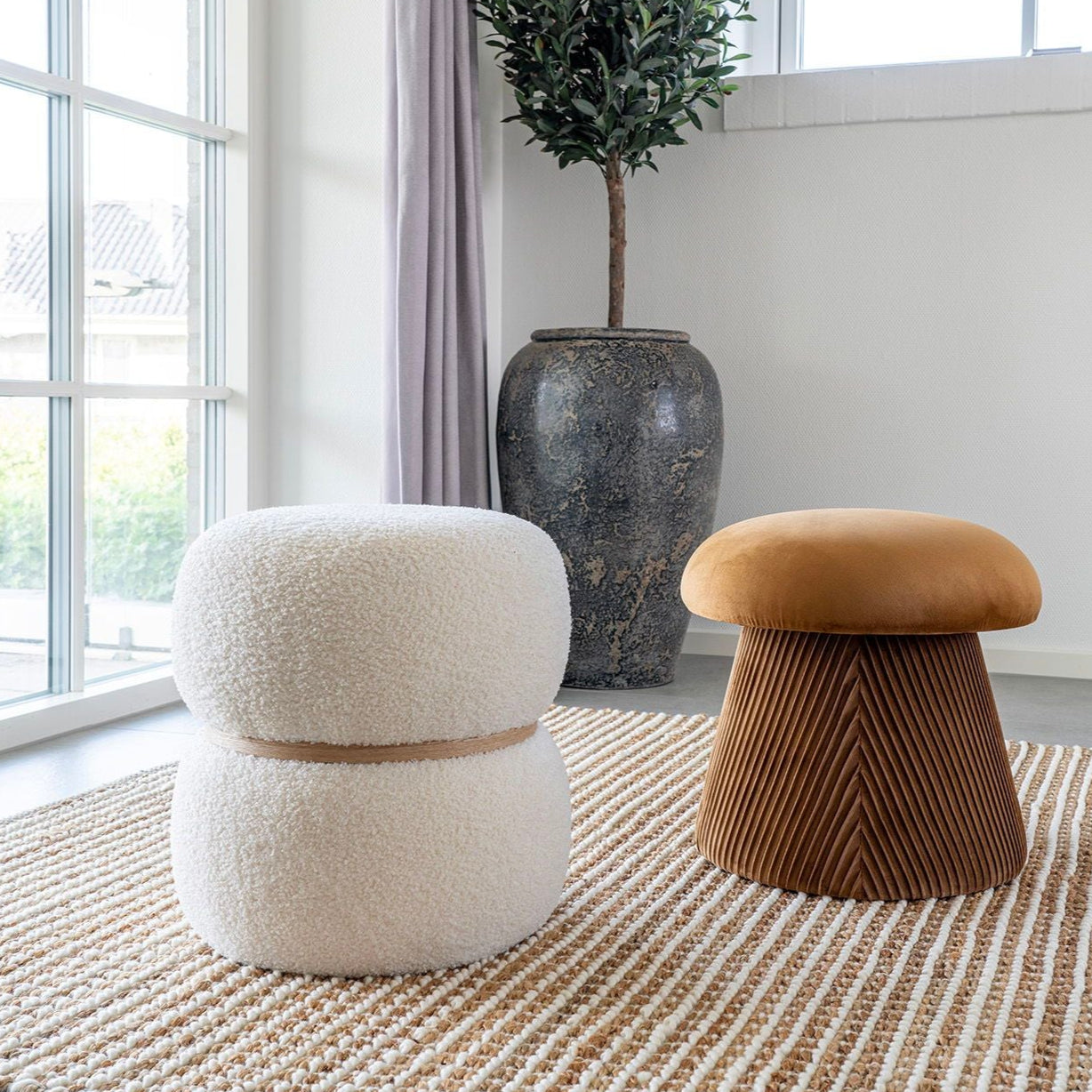 Ett elegant rum med sofistikerade detaljer med två pallar på en randig matta. Den ena är den eleganta Milford-puffen från House Nordic, vit och fluffig som lyxiga bouclé-kuddar. Den andra pallen är brun med en veckad bas. En hög växt i en stor vas står nära skira gardinfönster.