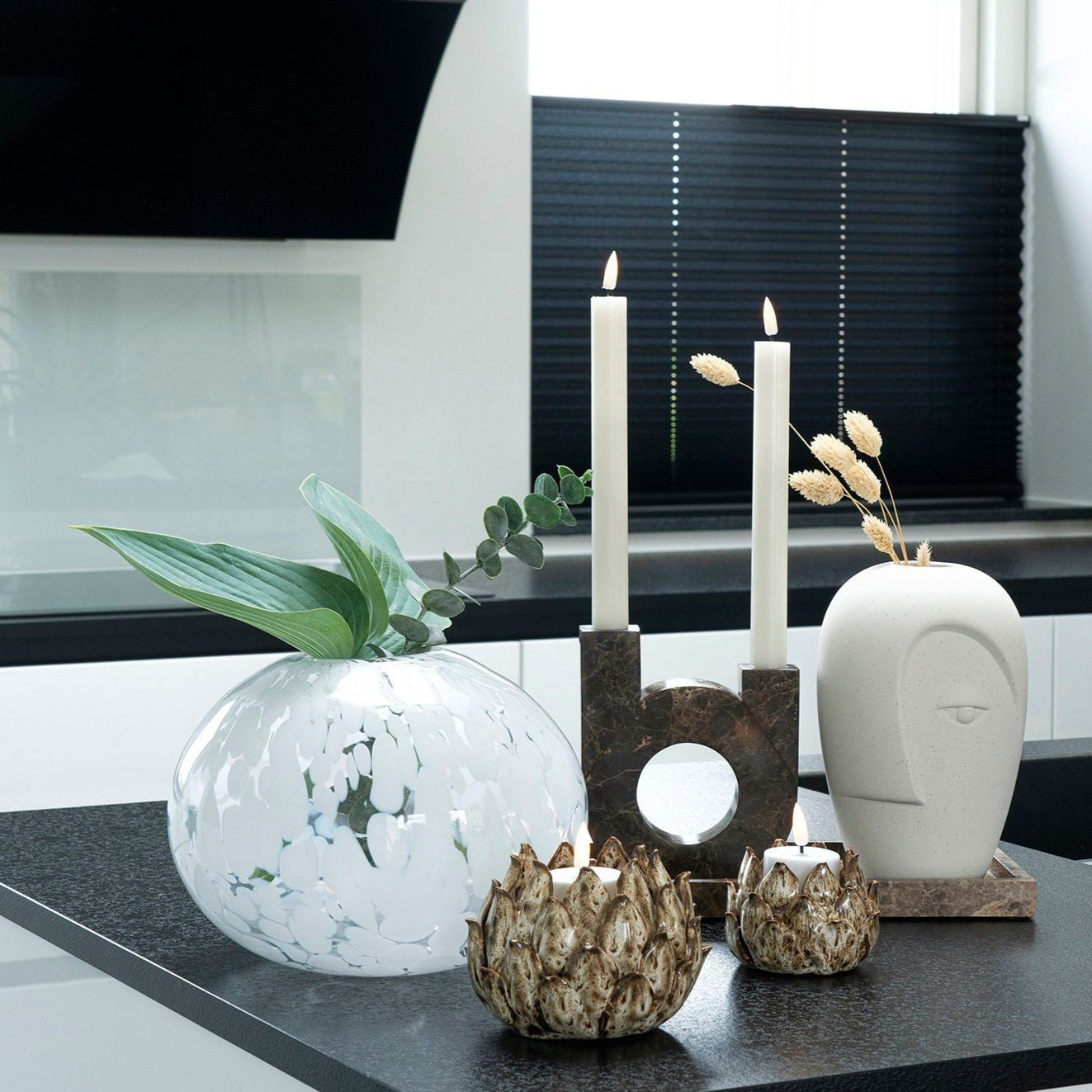 Ett modernt kök i U-format har en vit och svart färgpalett med en mörk bänkskiva. Den pryds av en dekorativ glasvas med gröna blad, två upplysta House Nordic U-formad ljusstake i brun marmor i abstrakta hållare och en hög keramisk huvudskulptur.