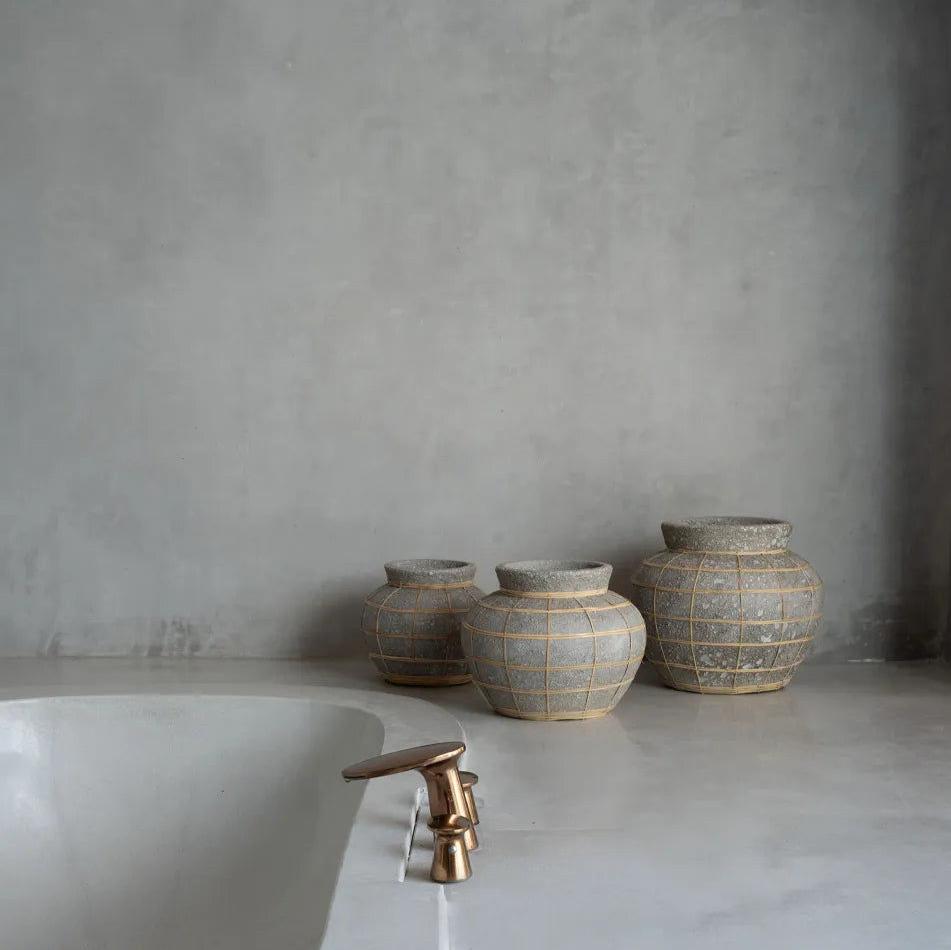 En minimalistisk badrumsscen visar upp en elegant, modern kran över ett vitt handfat till vänster, medan tre Terracotta Vas med Rattandetalj från bazar bizar med rottingdetaljer lägger till textur till höger, allt mot en slät, ljusgrå bänkskiva och vägg.