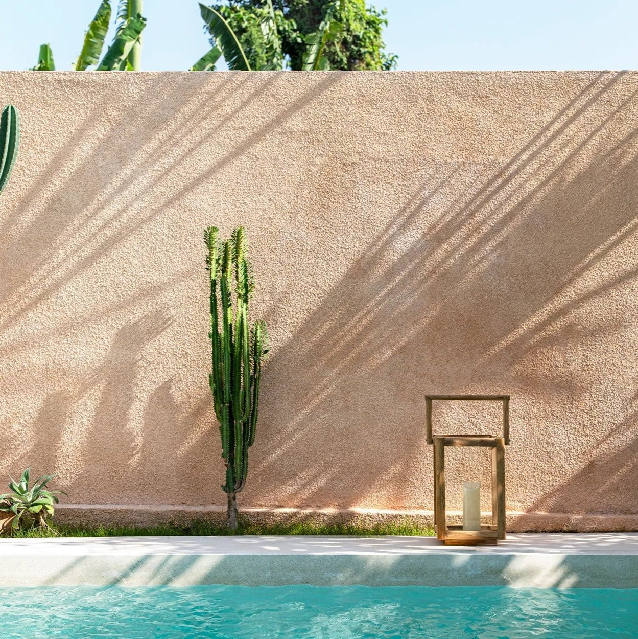 En lugn utomhusscen har en texturerad beige vägg med växtskuggor, en grön kaktus vid väggen och en turkos pool under en klar himmel. Den eleganta Nusa Penida Lykta - Small by bazar bizar vilar på marken bredvid poolen.