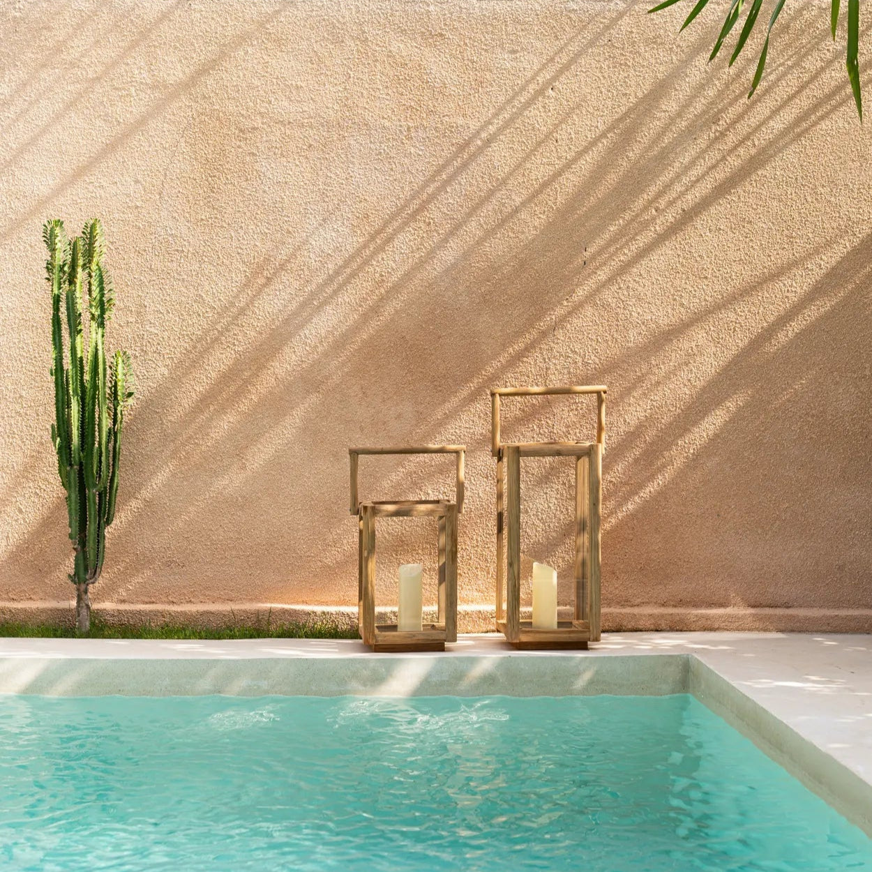 En turkos swimmingpool bredvid en beige vägg, hög kaktus och två Nusa Penida Lykta från Bazar Bizar i hållbart teakträ skapar en harmonisk ljusmiljö för din uteplats.