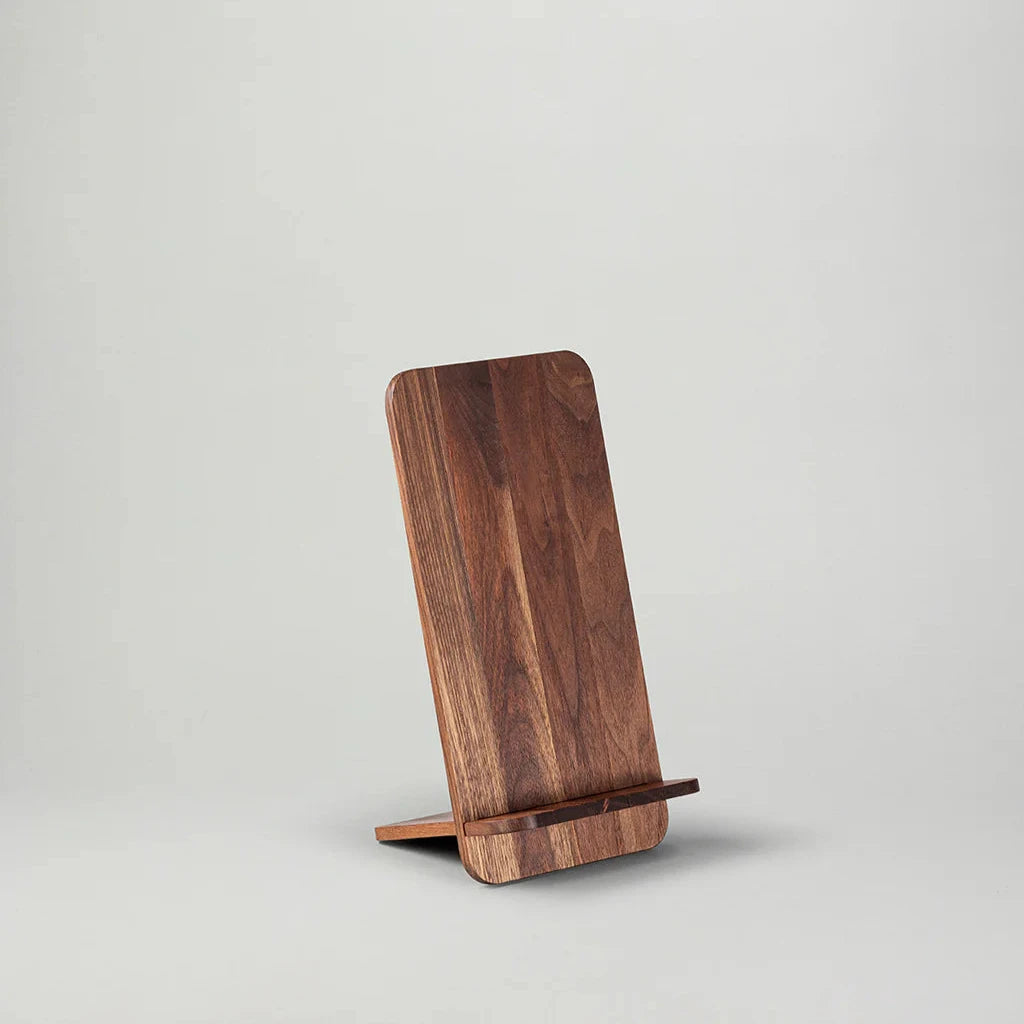 Vi presenterar NM Bokställ – Valnöt av New Mags, en elegant surfplatta eller telefonstativ i mörk trä i minimalistisk design. Stativet, prydt med en valnötsfinish, inkluderar ett lätt vinklat ryggstöd och en liten avsats i botten för att säkert hålla din enhet. Bakgrunden är vanlig ljusgrå.