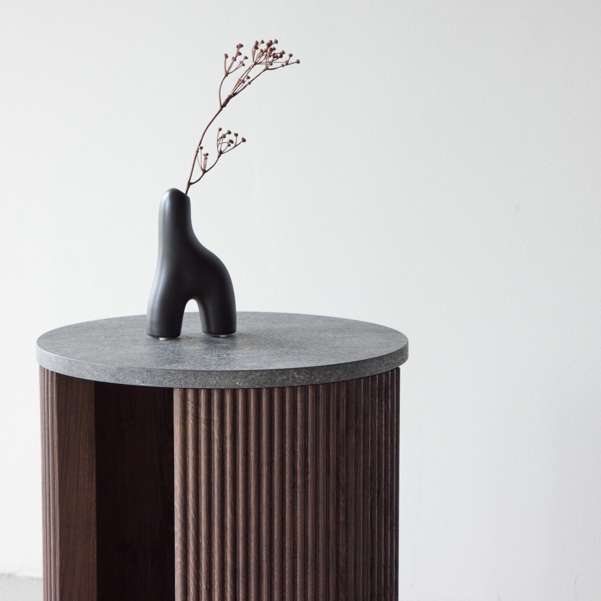 En minimalistisk mörk vas med torkade grenar står på Venture Homes Bristol Sidobord - Rund marmorlook i mocca, mot en vit vägg.