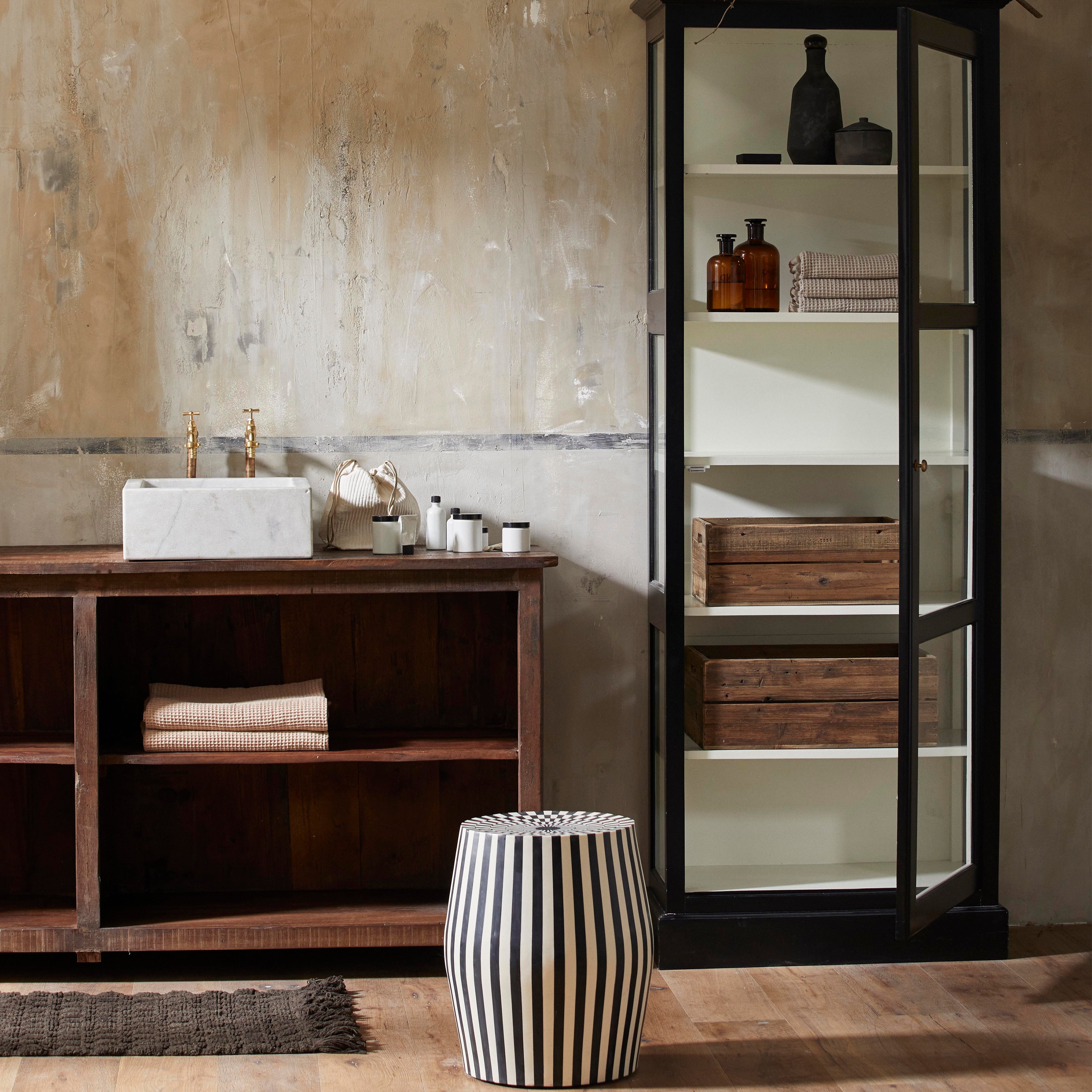 Ett rustikt badrum har ett kommod i trä, ett handfat i marmor och öppna hyllor. Nordal TAURA Sidobord – Konstnärlig Design i Svart och Ivory – ger trägolvet stil och framhäver handgjorda-materialet i hela utrymmet.
