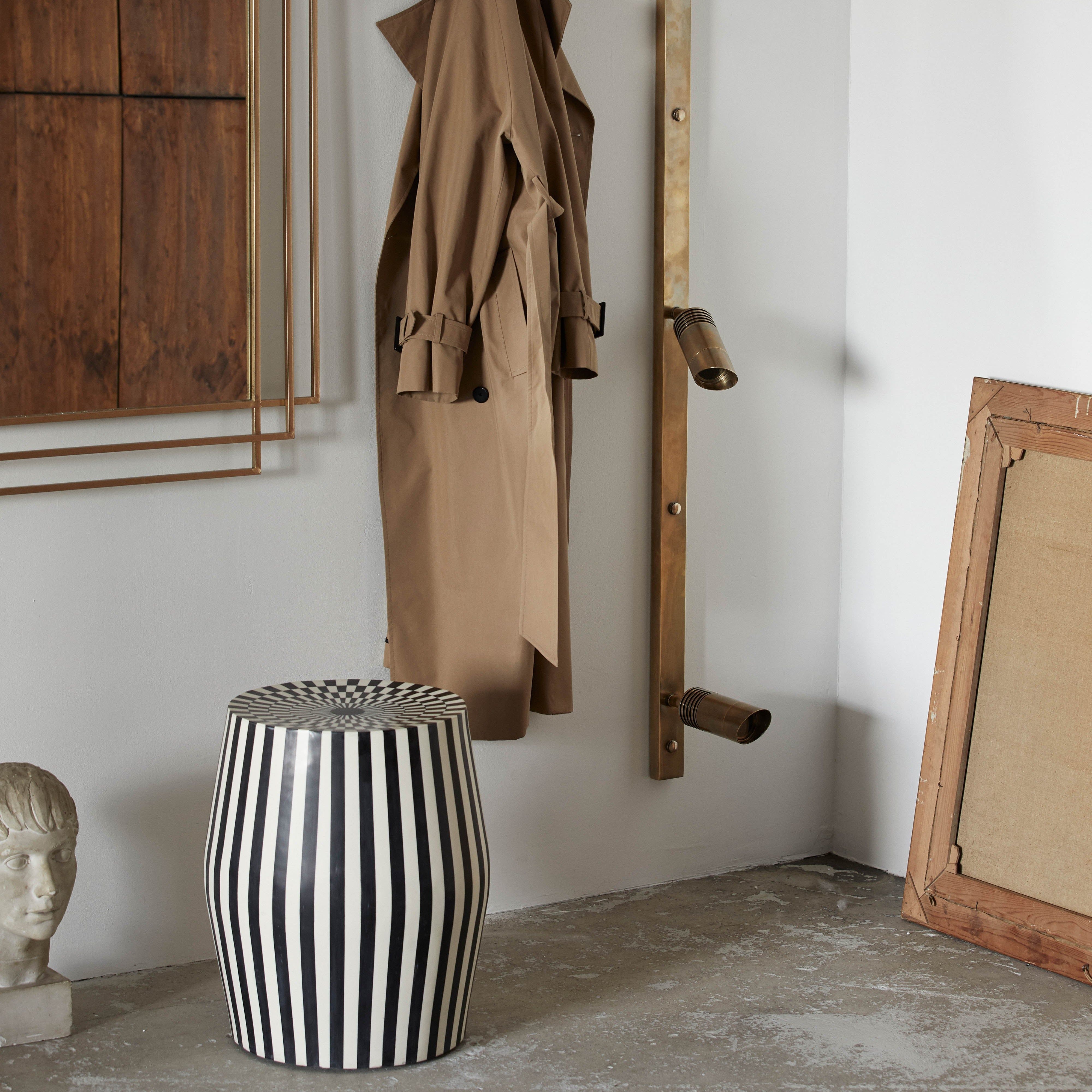 En brun trenchcoat hänger på ett väggställ i mässing bredvid en stor spegel, TAURA Sidobord – Konstnärlig Design i Svart och Ivory av Nordal, en randig svartvit pall, inramad duk och en delvis huvudskulptur på betonggolvet.