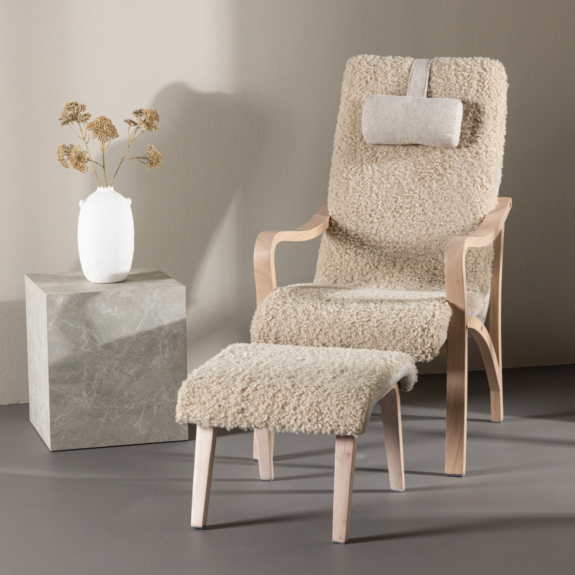 Den mysiga Fårö fåtölj med fotpall och nackkudde från Venture Home, i krämfärgad pälsliknande polyestertyg med träben, andas nordisk elegans mot en neutral vägg. En vit vas med torkade blommor vilar på en grå marmorsockel i närheten.