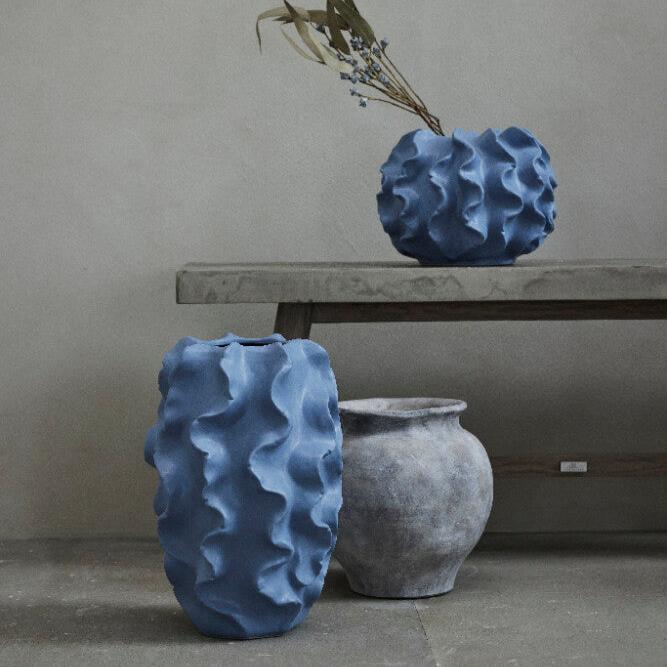 En strukturerad blå vas med en vågig yta sitter på en minimalistisk bänk och rymmer några torkade blommor. Under den, på golvet, finns ytterligare två vaser: en matchar den blå vasens struktur och nyans, medan den andra är en elegant Anna Handgjord Vas i Antikgrå H26,5 cm från Lene Bjerre Design DK—klassisk tidlös design.