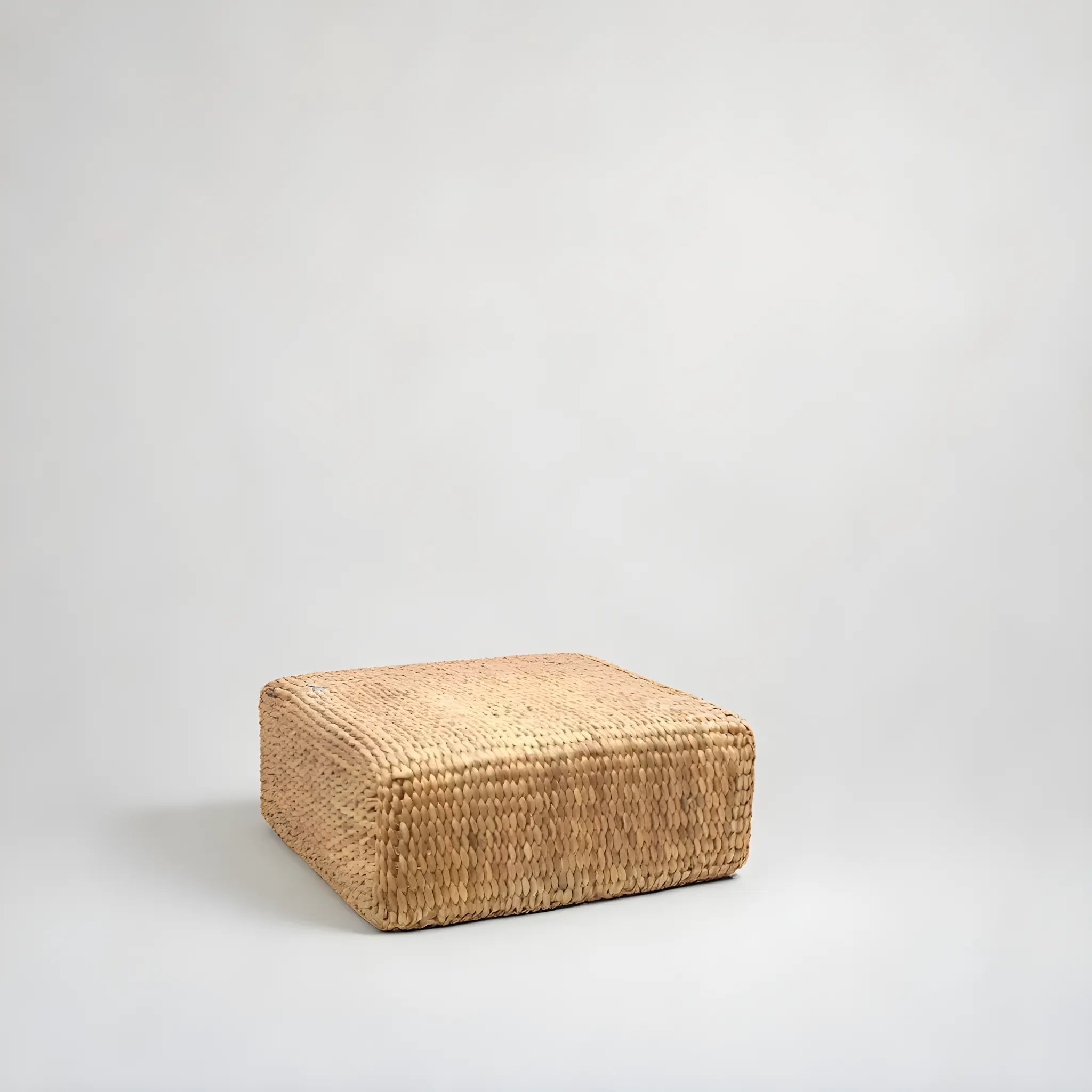 Fyrkantig Puff i Vattenhyacint av bazar bizar vilar på en ljusgrå bakgrund, med en strukturerad yta i naturlig beige. Dess minimalistiska och rustika design gör den perfekt för inredning, och erbjuder osmyckad enkelhet.